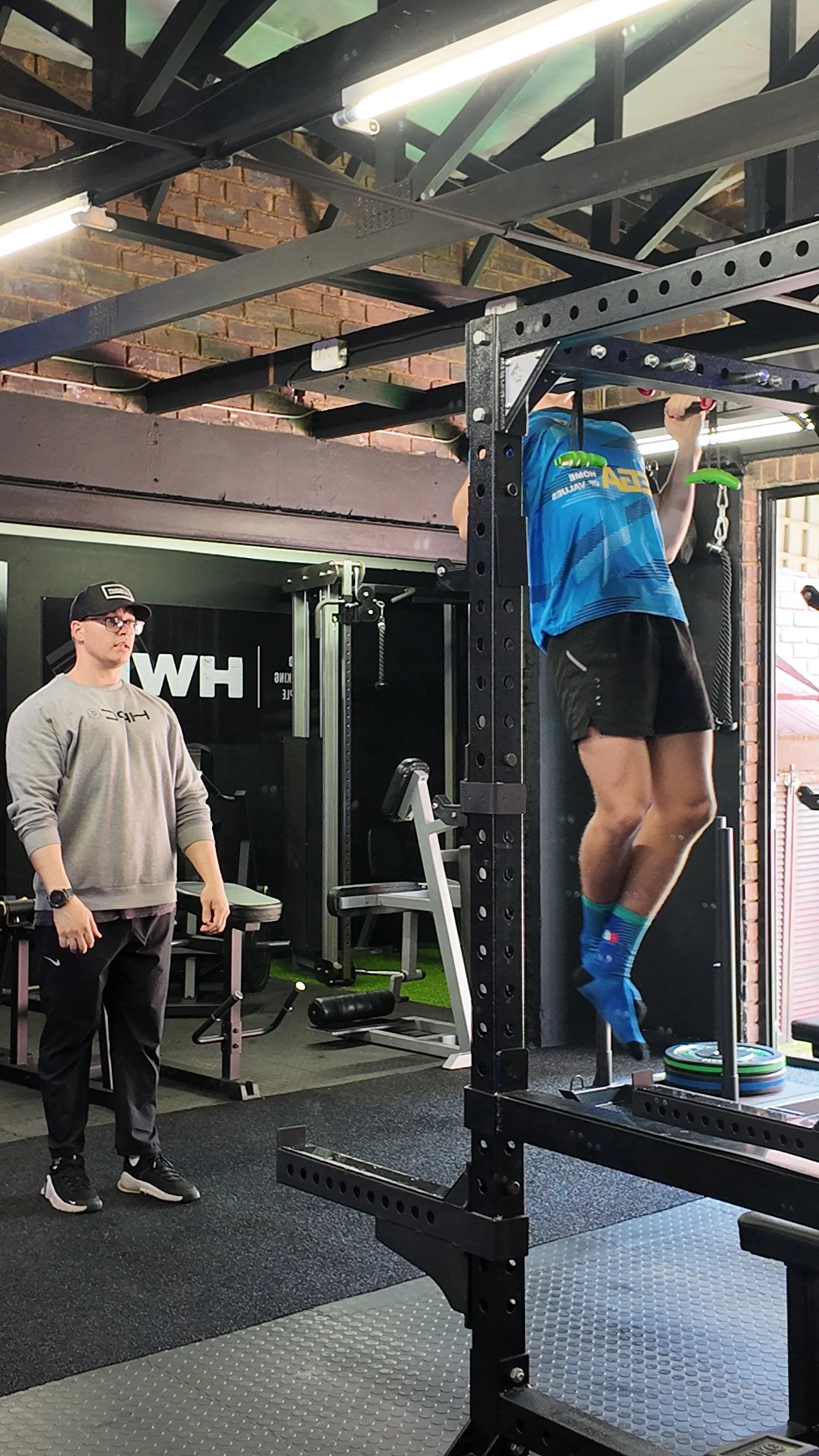 A person is performing a pull-up exercise on a pull-up bar in a gym, while another person stands nearby watching. The gym has brick walls and workout equipment.