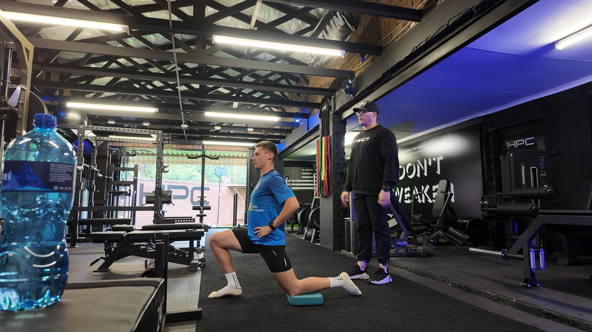 A man stretching on one knee during a workout in a gym, with a trainer standing nearby. There is gym equipment, a large water bottle, and motivational signage on the wall.