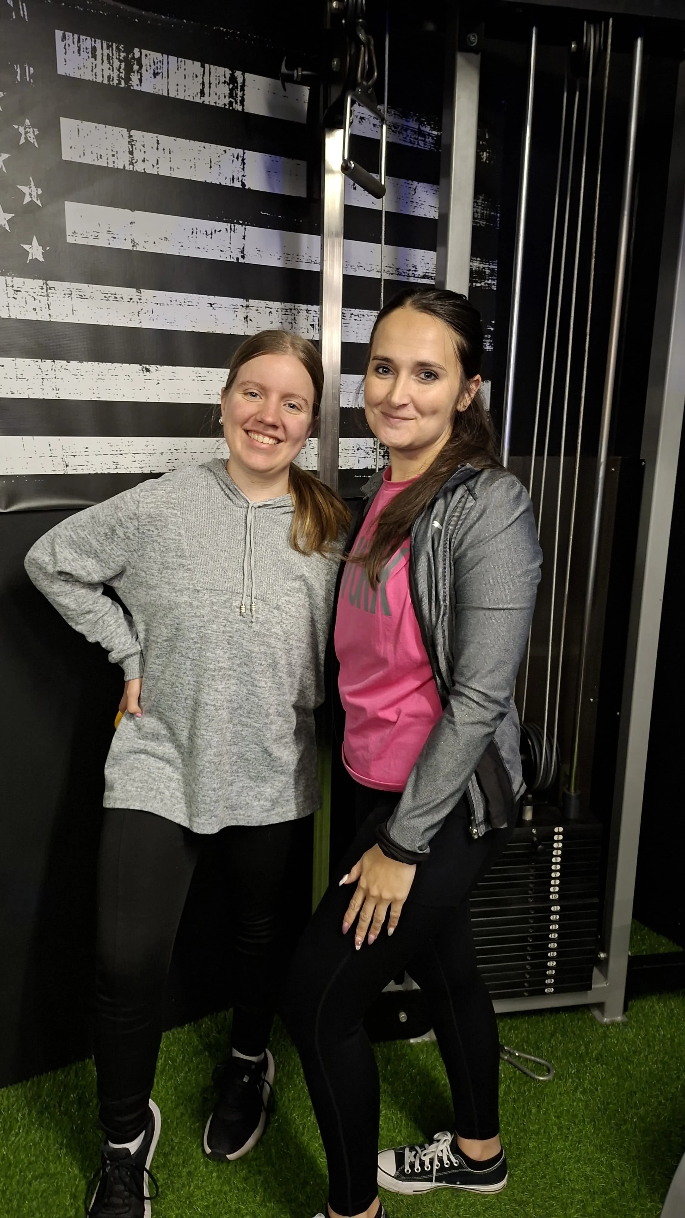 Monique Labuschagne and Geneveve smiling and posing in front of a large American flag graphic and exercise equipment at the HPC gym, wearing workout clothes.