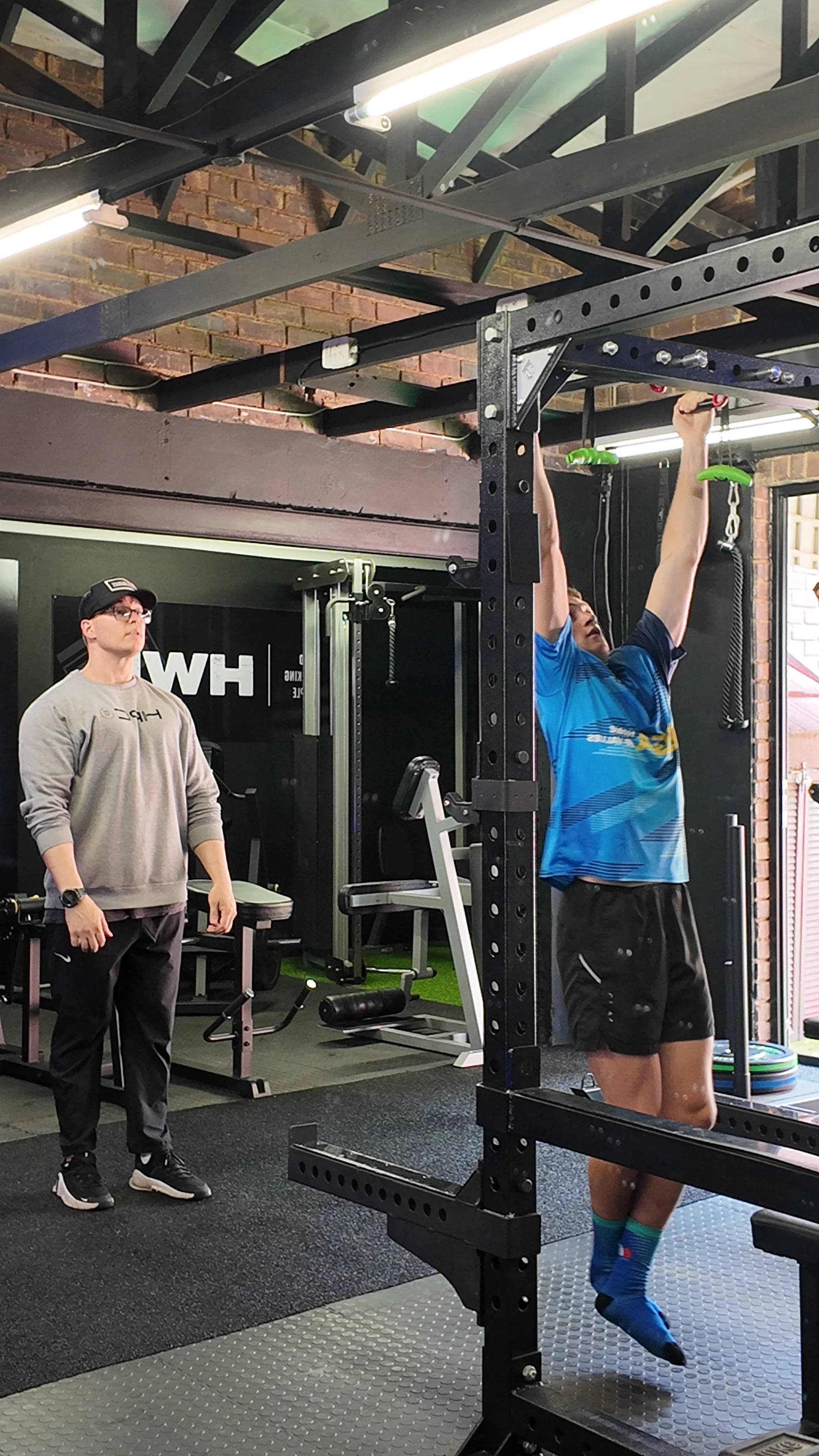 A man in a blue shirt doing pull-ups on a pull-up bar in a gym, with another man observing nearby.