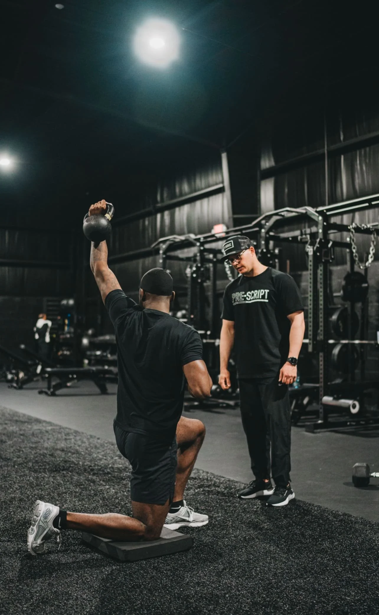 A man is kneeling on one knee, holding a kettlebell in his right hand in a gym. Another man is standing in front of him, wearing a black t-shirt with the word 'PRE-SCRIPT' and a black cap. The gym has black equipment and dark walls.
