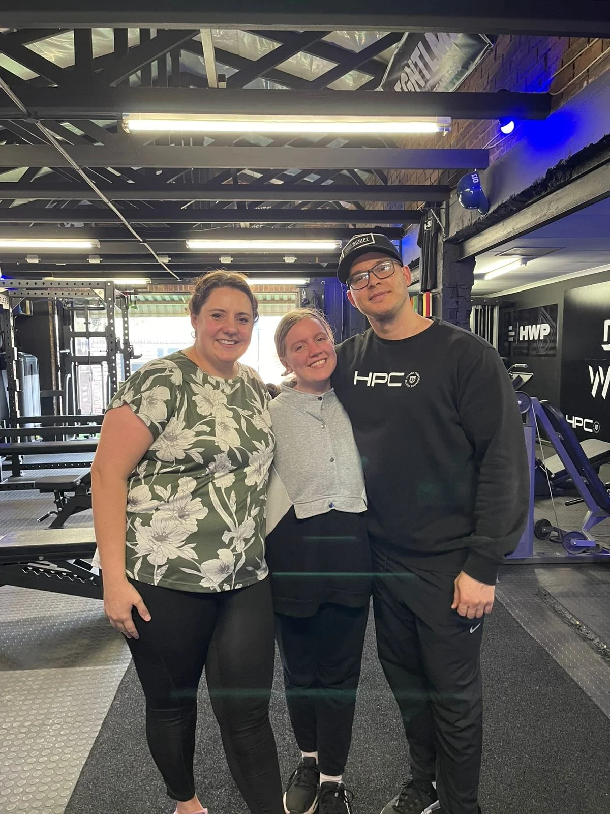 Anton Esterhuizen, Monique Labuschagne and Corlian smiling at the camera inside the HPC gym with workout equipment and black walls.