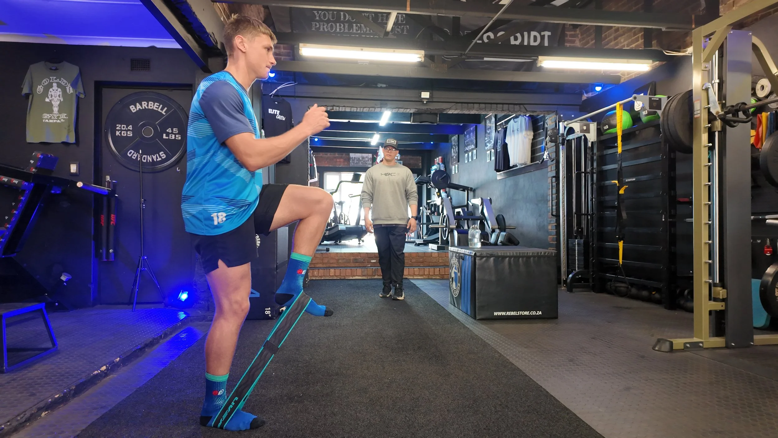 A young man wearing a blue sports jersey and shorts is performing a fitness exercise with resistance bands in a gym. A man in a gray hoodie and cap is observing in the background. The gym has various exercise equipment, weights, and T-shirts on displ