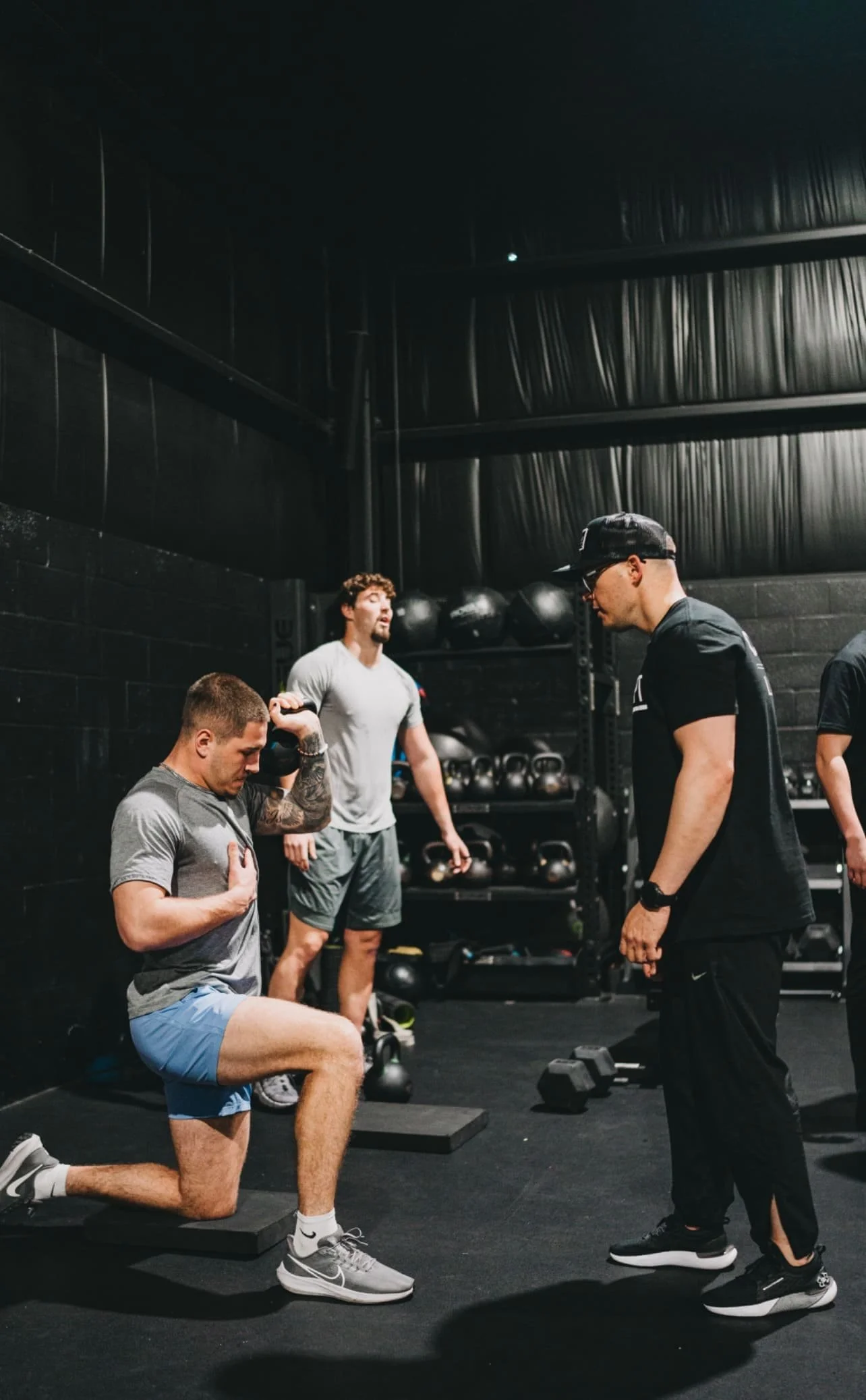 A man kneeling on a black exercise mat performing a knee push-up with one hand on his heart, in a gym with black walls and shelves of kettlebells and medicine balls. Three other men are standing nearby, two of them watching and one with a camera in hand.