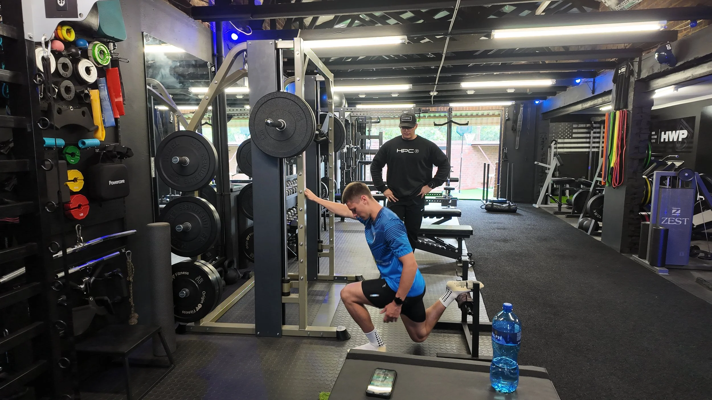 A gym with two men, one sitting and one standing, engaging in workout activities. The gym has various weights, resistance bands, and exercise equipment visible. There is a water bottle on a small black table in the foreground.