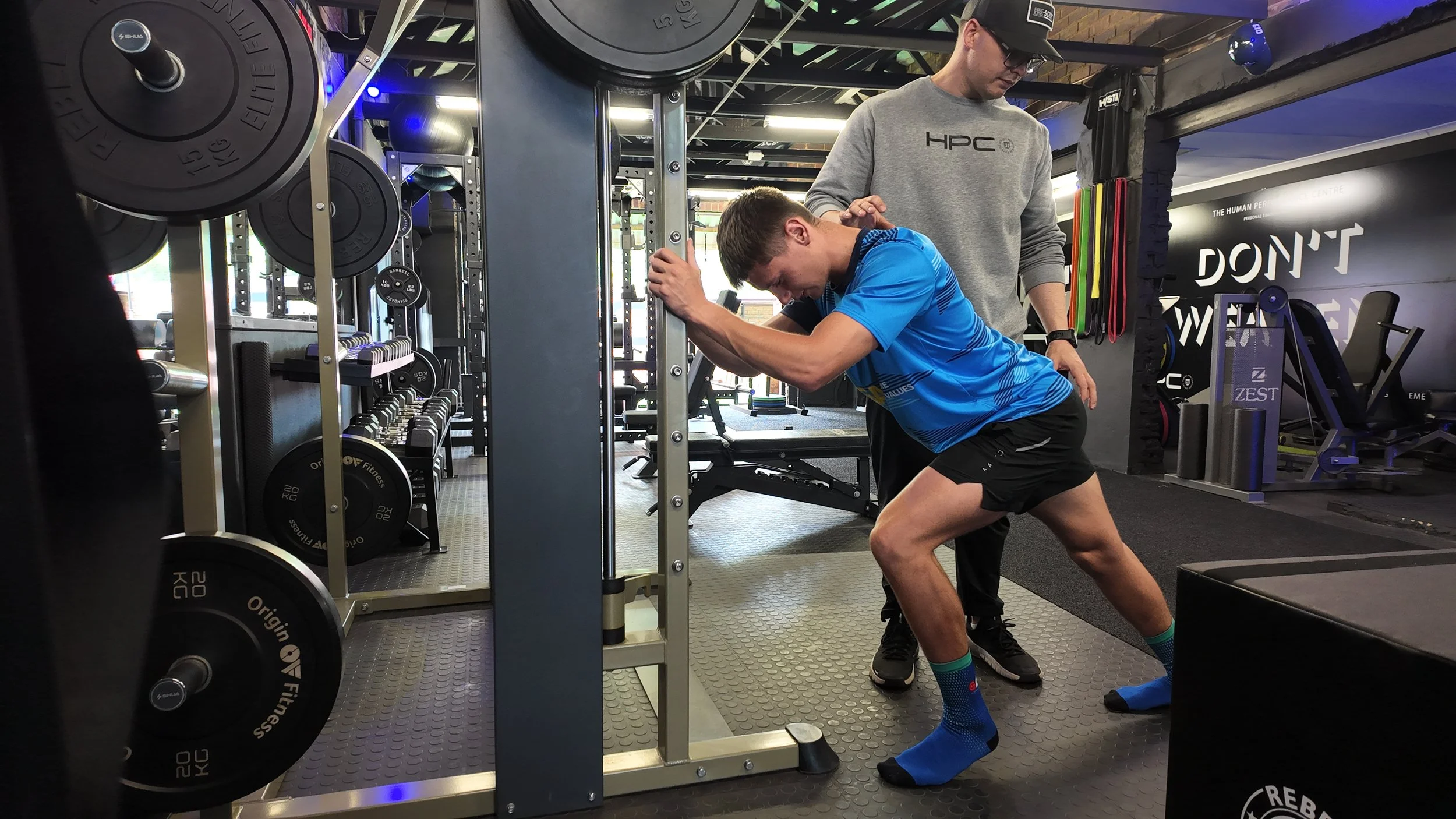 A man in a blue shirt and black shorts performing a squat exercise with a spotter in a gym. The gym has weight plates, exercise equipment, and motivational posters.