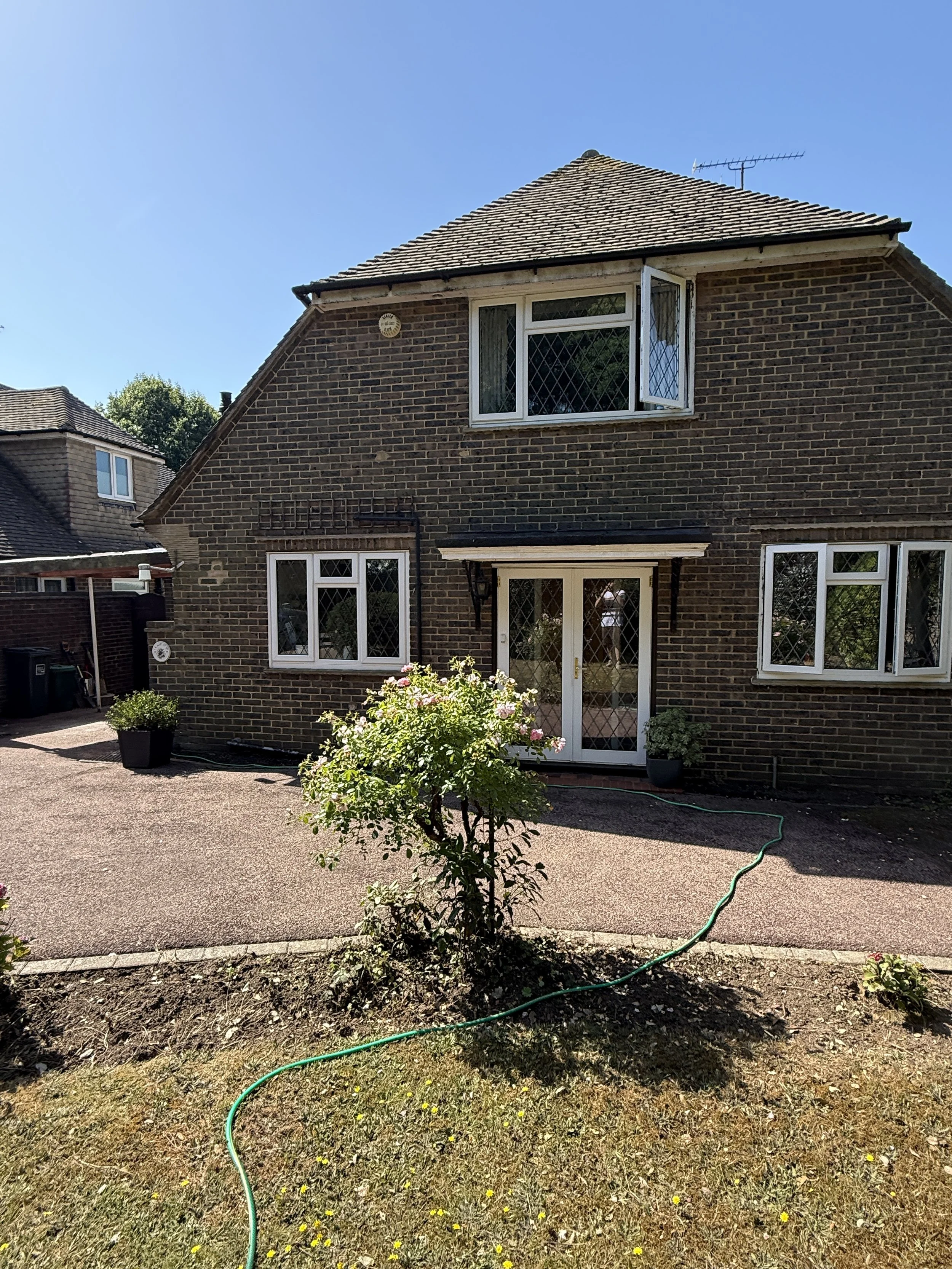 A brick house with a sloped roof and white-framed windows, a garden with a small tree and flowering bushes, and a green garden hose laid on dry grass in the foreground under a clear blue sky.