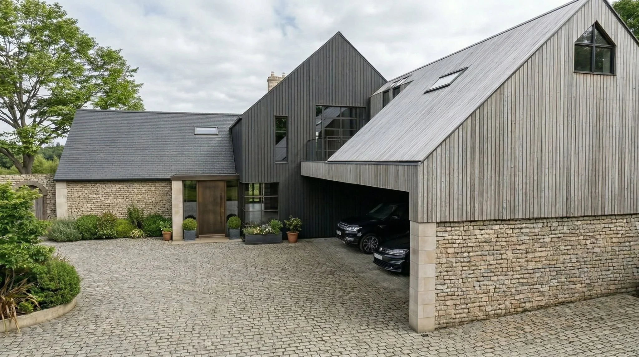 Modern house with a combination of stone and black wood siding, large windows, and a garage with two cars parked underneath. There is a paved driveway with decorative plants near the entrance, and a tree on the left side of the house.