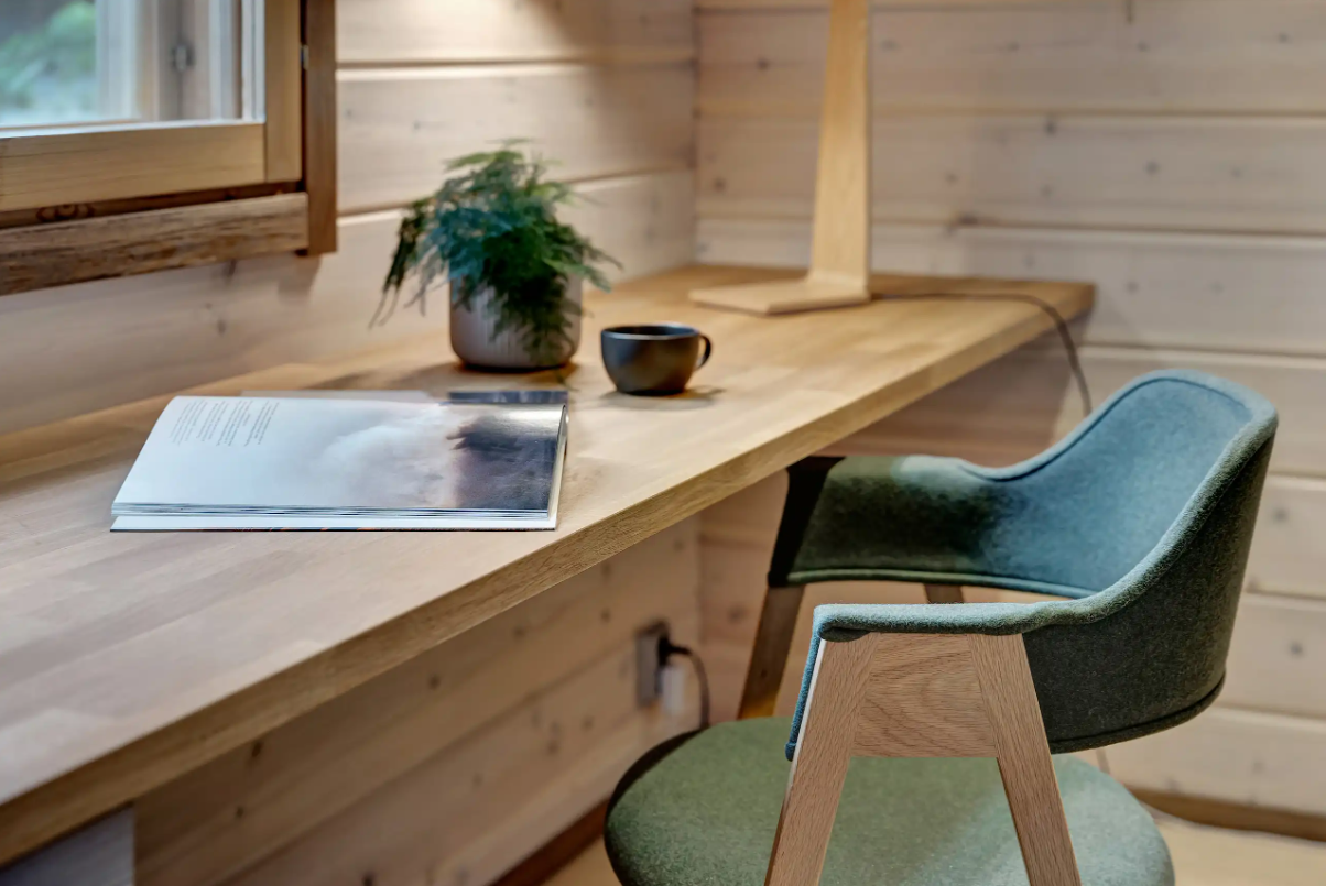 Ein Holzschreibtisch mit einer offenen Zeitschrift, einer schwarzen Tasse, einer Pflanze in einem Topf und einem Laptop. Daneben ein bequemer Schreibtischstuhl mit grüner Rückenlehne in einem Raum mit Holzvertäfelung.