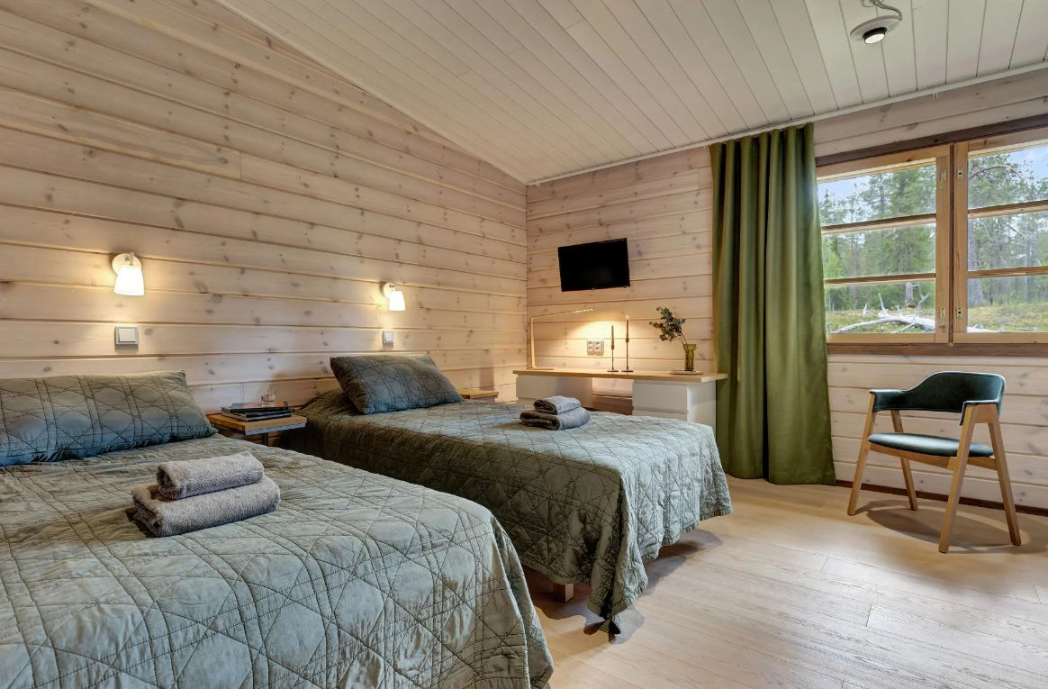 Ein Schlafzimmer mit zwei Betten in einem Holzhaus, einem kleinen Fernseher an der Wand, eine Lampe auf einem Schreibtisch, ein Fenster mit grünen Vorhängen und einem Blick in eine Naturlandschaft.