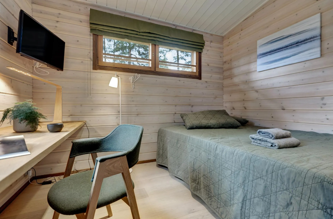 Ein Schlafzimmer mit Holzverkleidung, einem Bett mit grünner Decke, einem Fenster, Wandkunst, einem Schreibtisch mit Stuhl, einer Lampe, einem Fernseher und einer Topfpflanze.