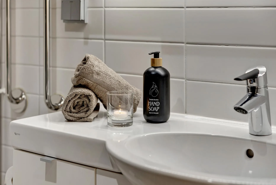 Waschbecken mit schwarzen Handseifenspender, zwei beige Handtücher, einer Kerze in einem Glas, weiße Fliesen an der Wand, Wasserhahn.