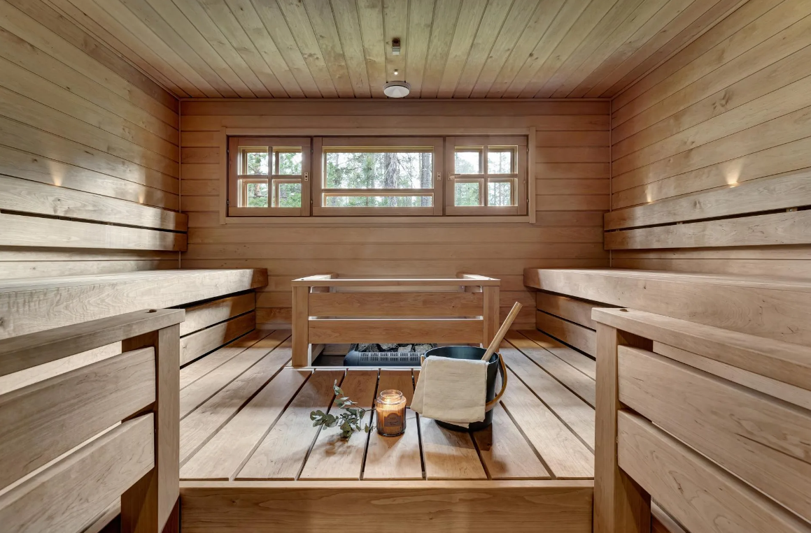 Innenraum einer traditionellen finnischen Sauna aus Holz mit Liegen an den Wänden, ein Fenster, ein Tisch, eine Kerze, ein Handtuch und ein Eimer mit Sauna-Utensilien.
