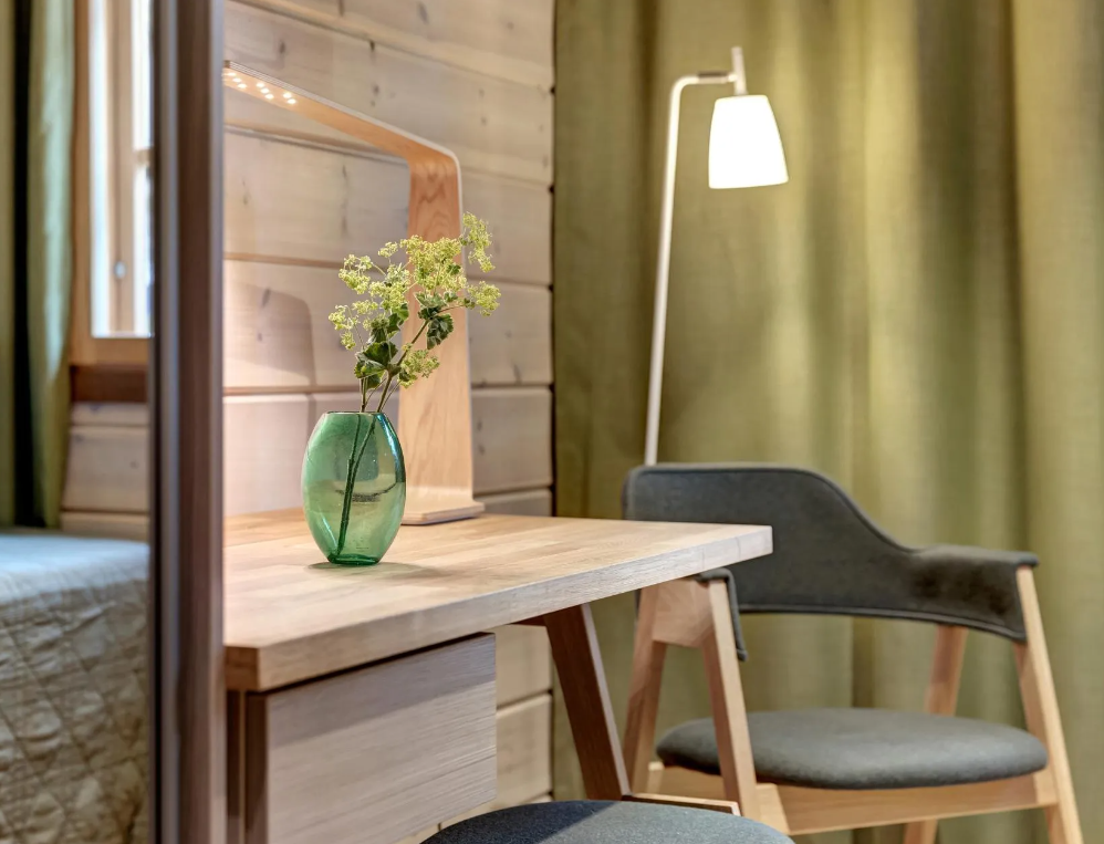 Ein Holzschreibtisch mit einer grünen Vase und Blumen, ein Fenster, ein grauer Stuhl und eine Stehlampe vor grünen Vorhängen in einem Raum mit Holzwand.