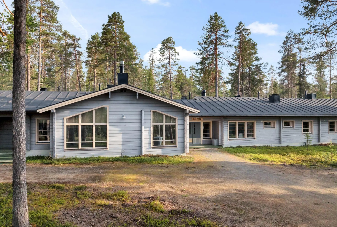 Ein modernes, graues Holzhaus in einem Waldgebiet mit vielen Tannenbäumen, unter einem blauen Himmel mit Wolken.