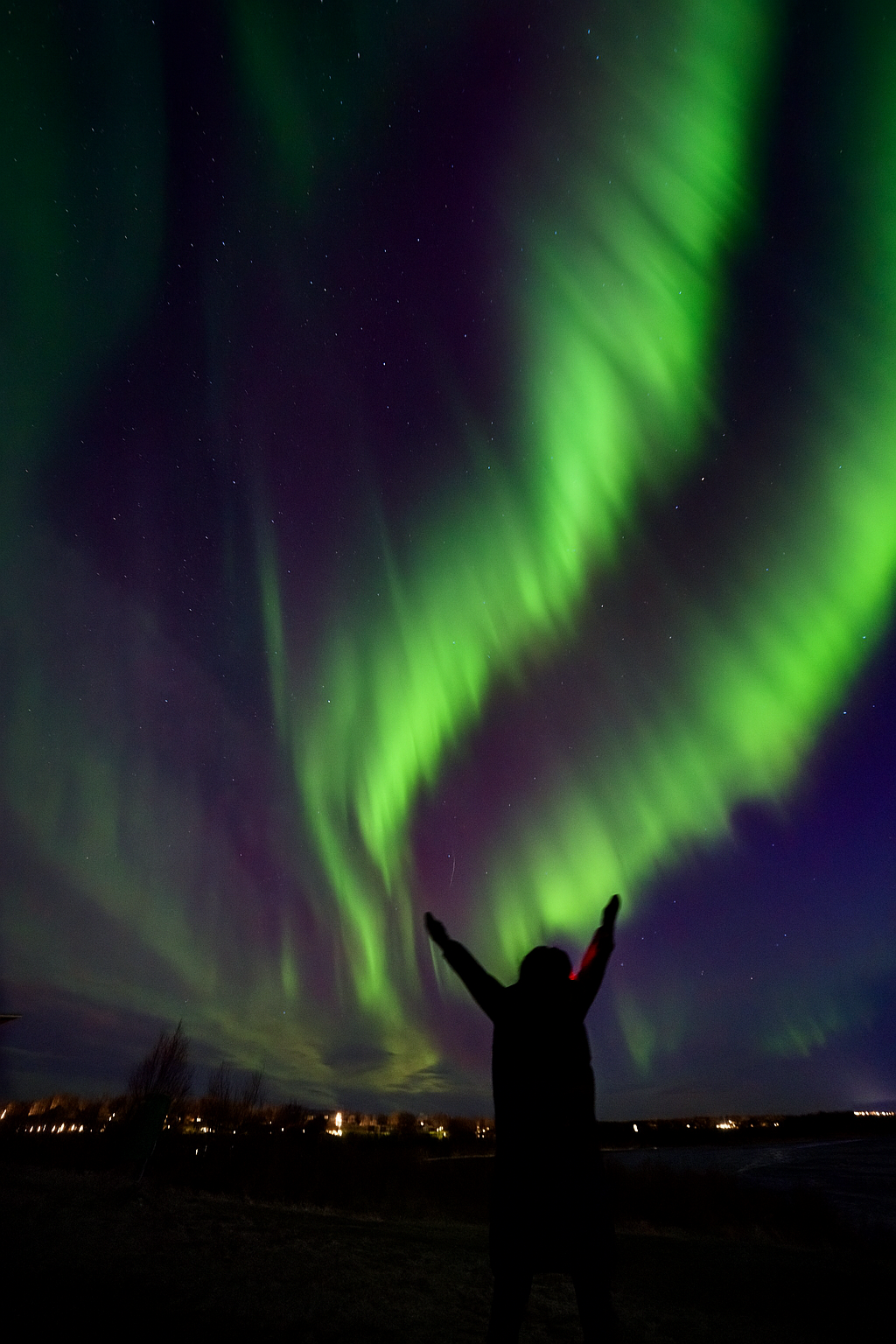 Eine Person steht im Freien bei Nacht mit erhobenen Armen, während die Nordlichter den Himmel mit grünen Lichtern erleuchten.