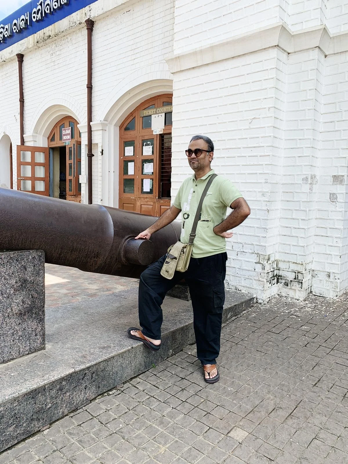 A man wearing sunglasses, a light green polo shirt, black pants, and sandals, standing outdoors beside a large historical cannon mounted on a stone platform, in front of a white brick building with arched windows and wooden doors, some signs and notices are posted on the doors.