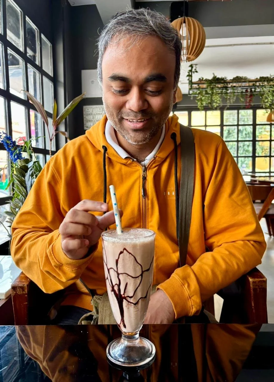 A man with gray hair, wearing a yellow jacket, sitting at a table in a cafe, stirring a chocolate milkshake topped with whipped cream and chocolate drizzle with a star-patterned straw.