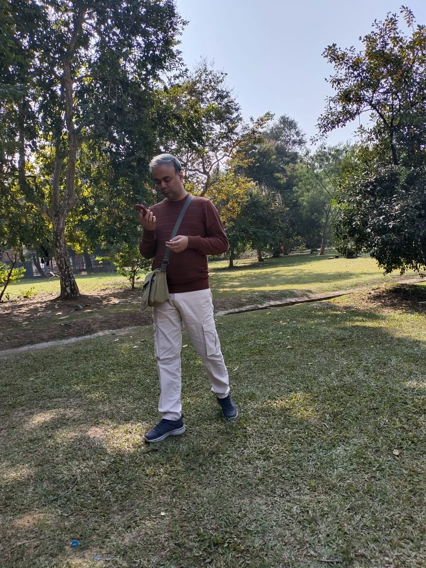 A man walking through a park while looking at his phone, dressed in a brown sweater, beige cargo pants, and black shoes, with a gray shoulder bag, surrounded by trees and grass.