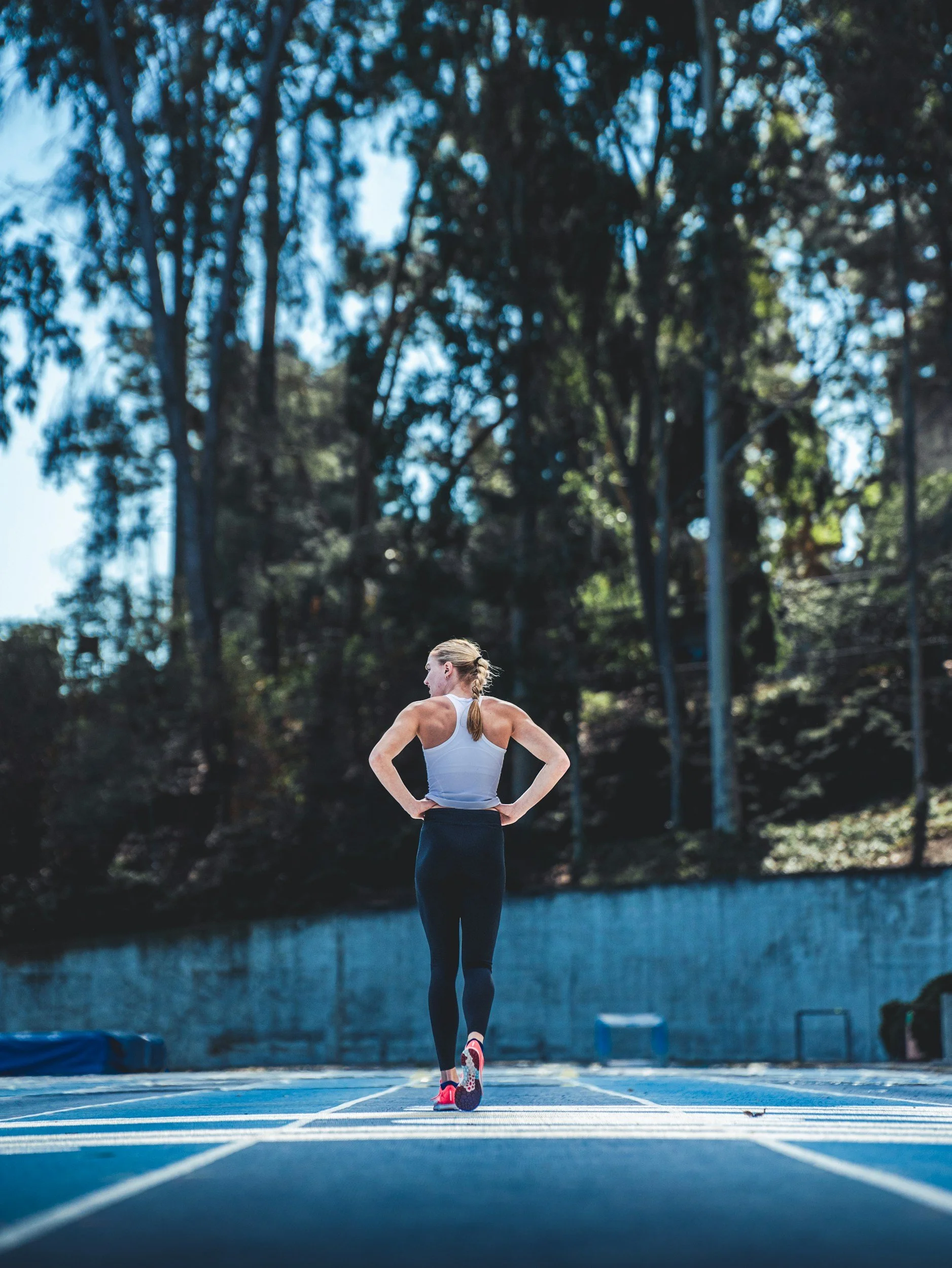 A woman in athletic wear standing on a running track, facing away with her hands on her hips, with trees in the background.