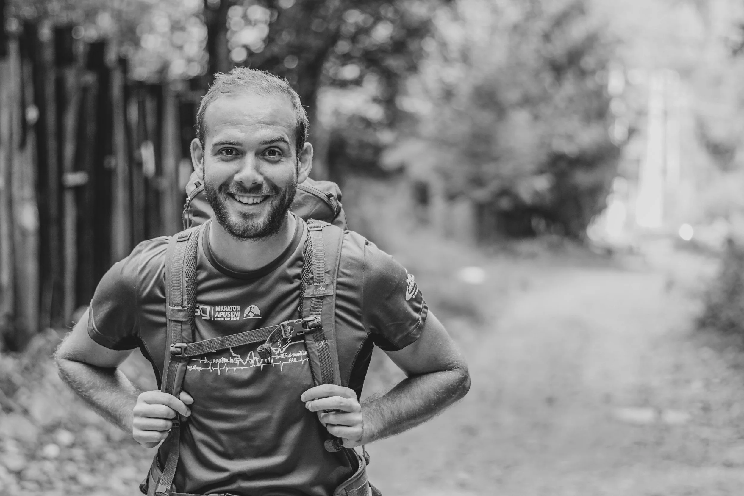 A man with a beard and short hair smiling while hiking outdoors, wearing a backpack and athletic clothing.