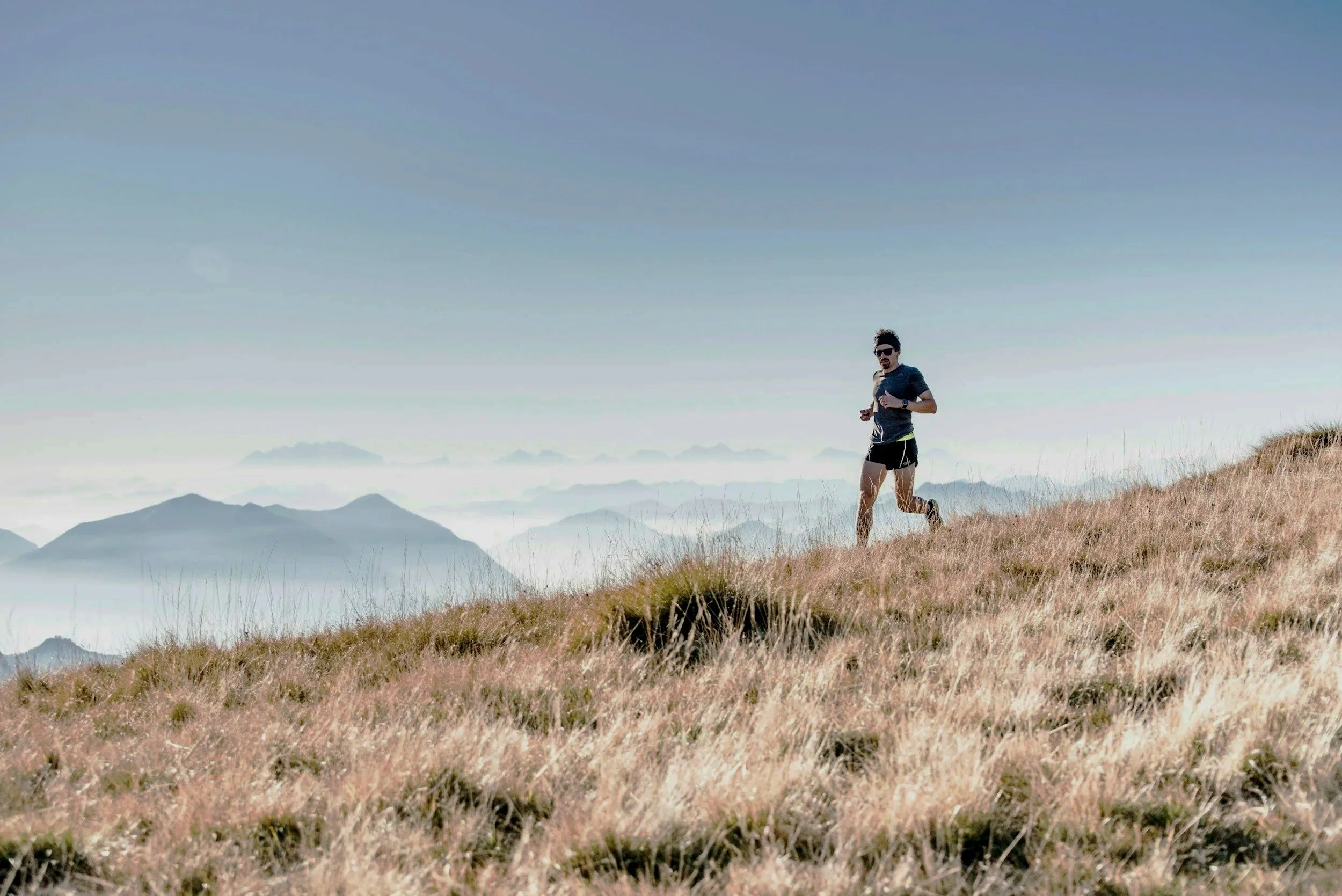 A man running on a grassy hillside with mountains and clouds in the background under a clear sky.
