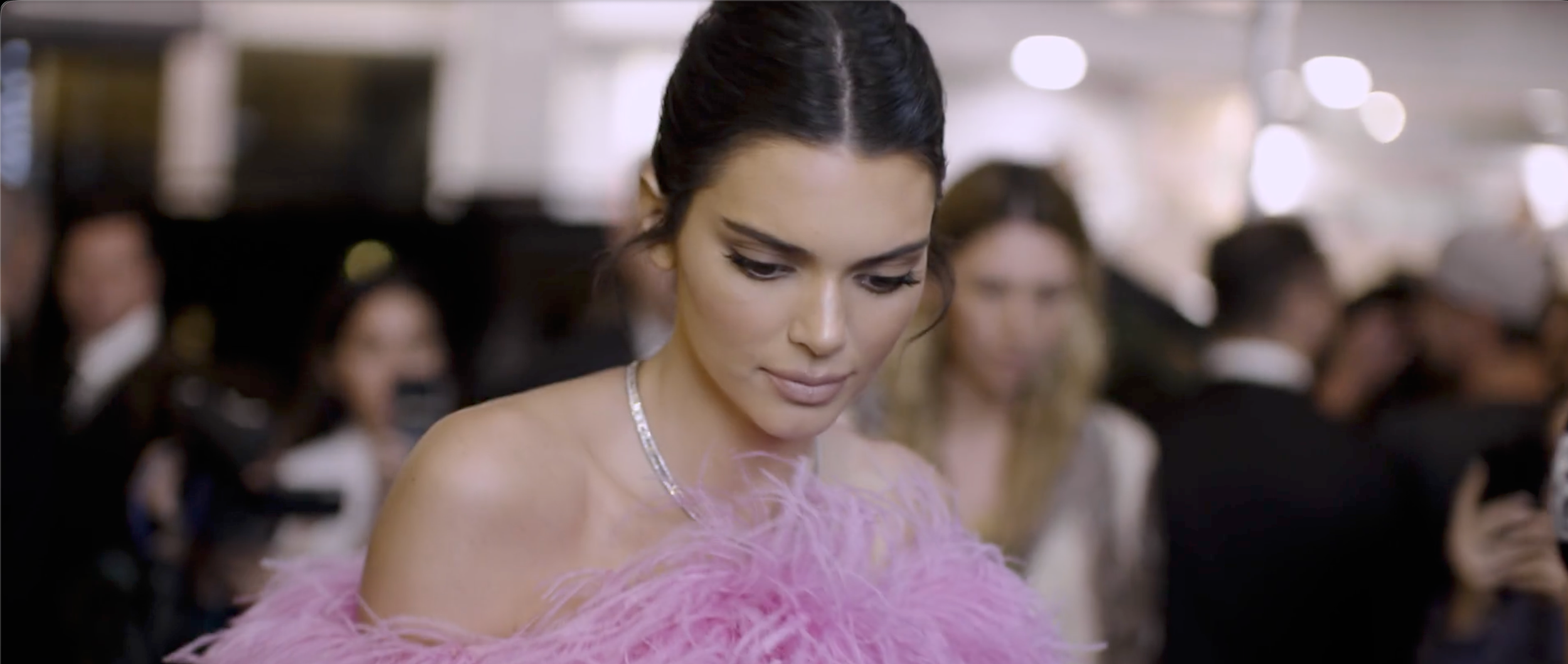 A woman in an elegant pink feathered dress at a formal event, with other guests blurred in the background.