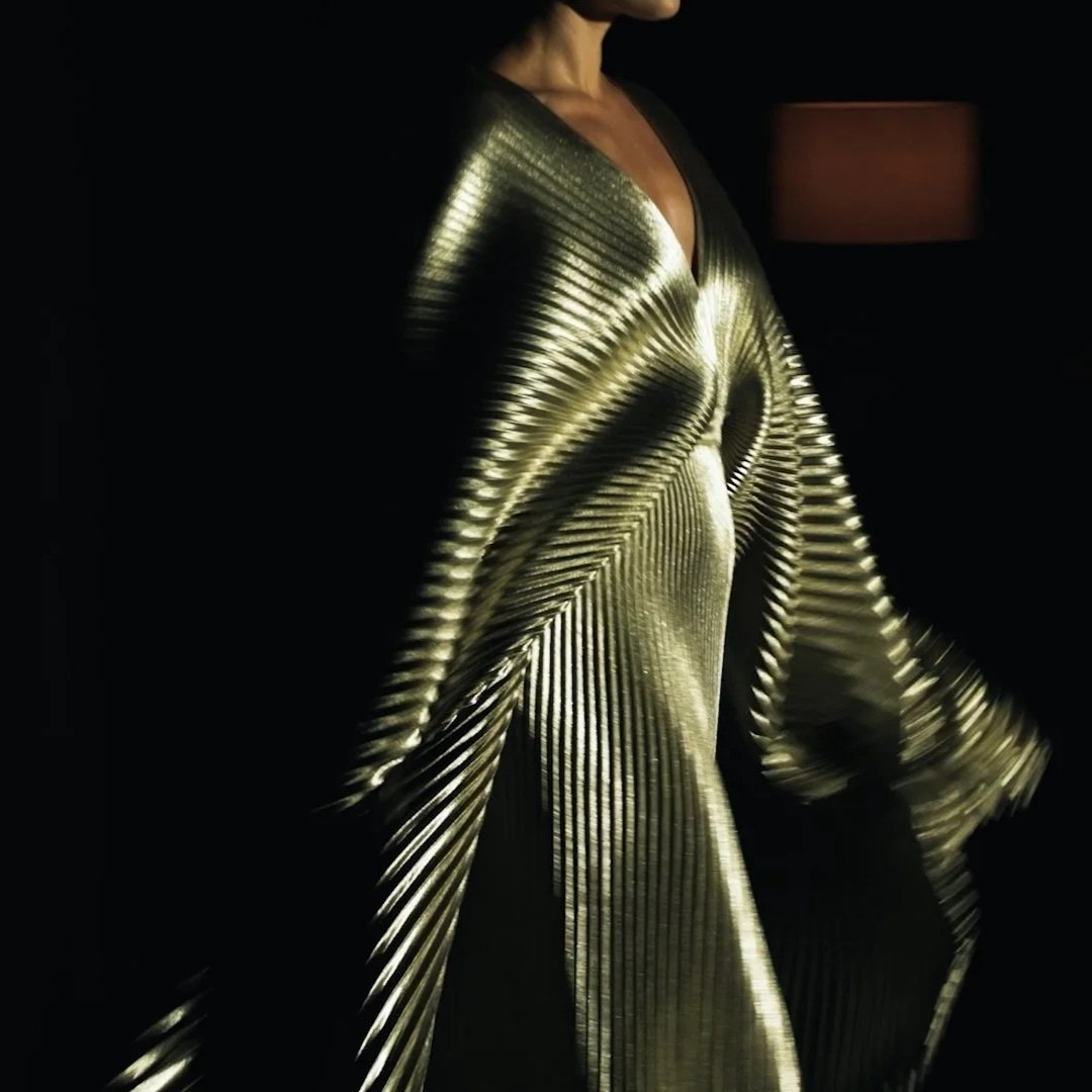 Person wearing a shiny, metallic gold dress with pleated design in a dark room, with a lamp in the background.