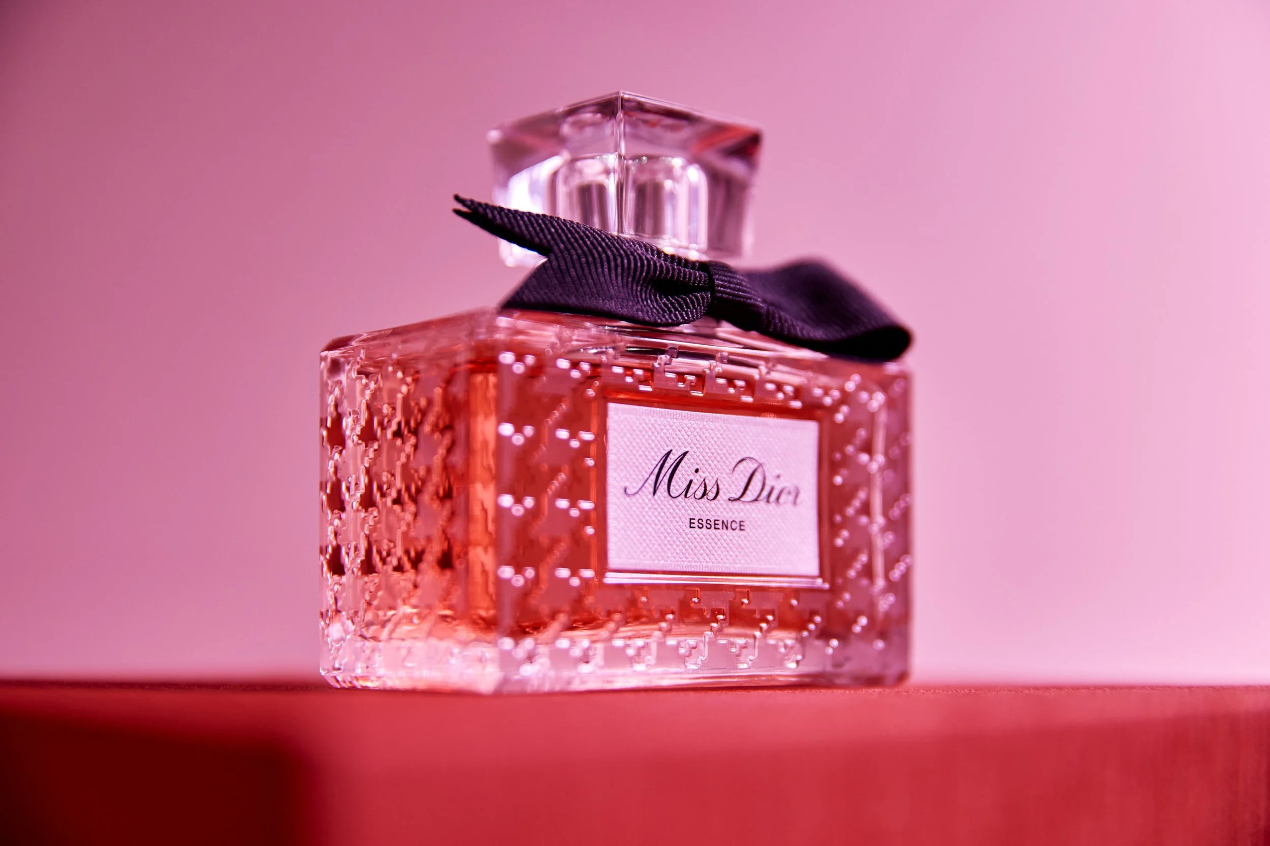 A pink perfume bottle with a textured pattern, labeled 'Miss Dior ESSENCE', topped with a clear square cap and a black ribbon, against a pink background.