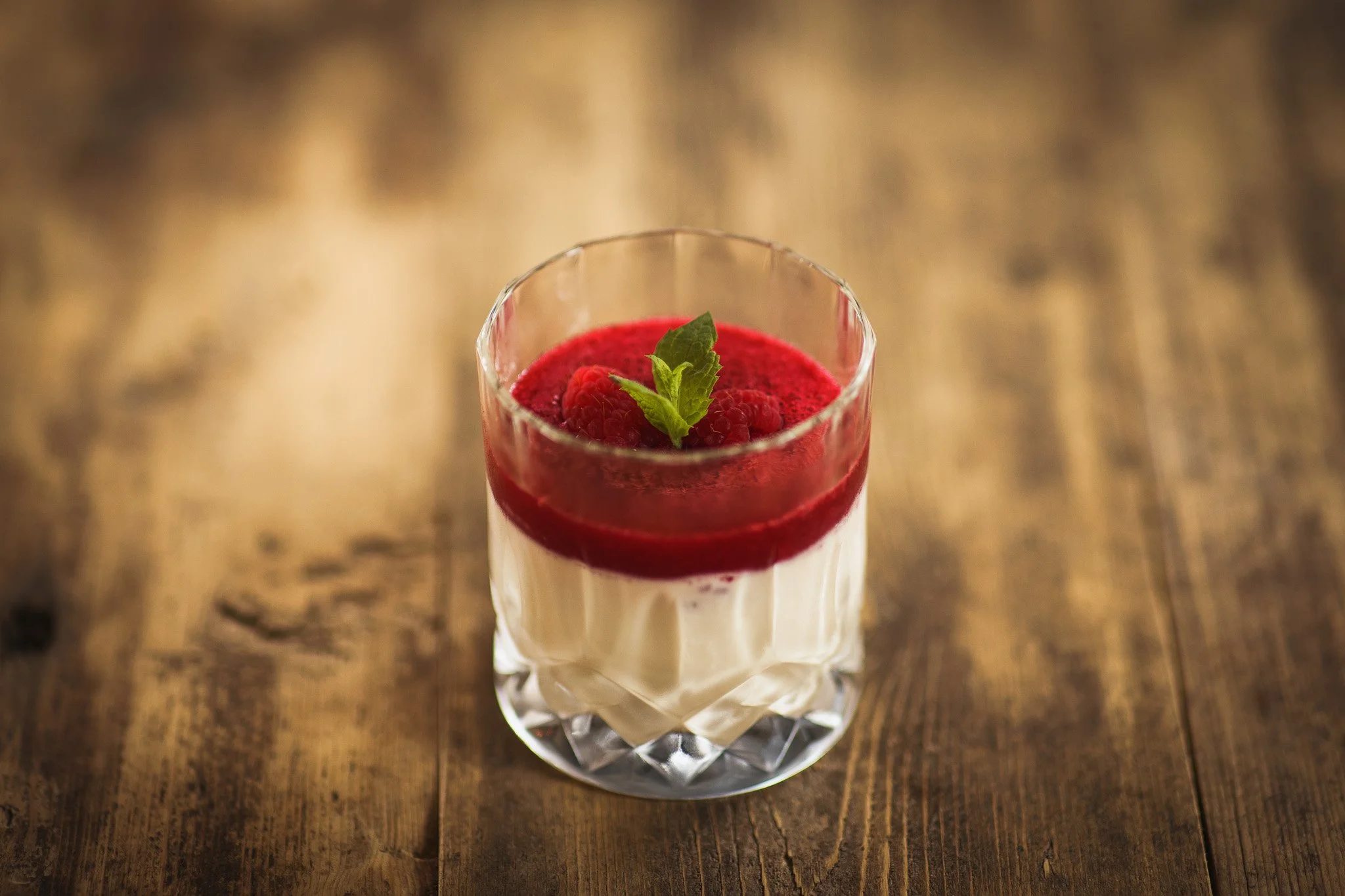 A dessert in a clear glass with a layered appearance, topped with raspberries and a mint leaf, placed on a wooden surface.