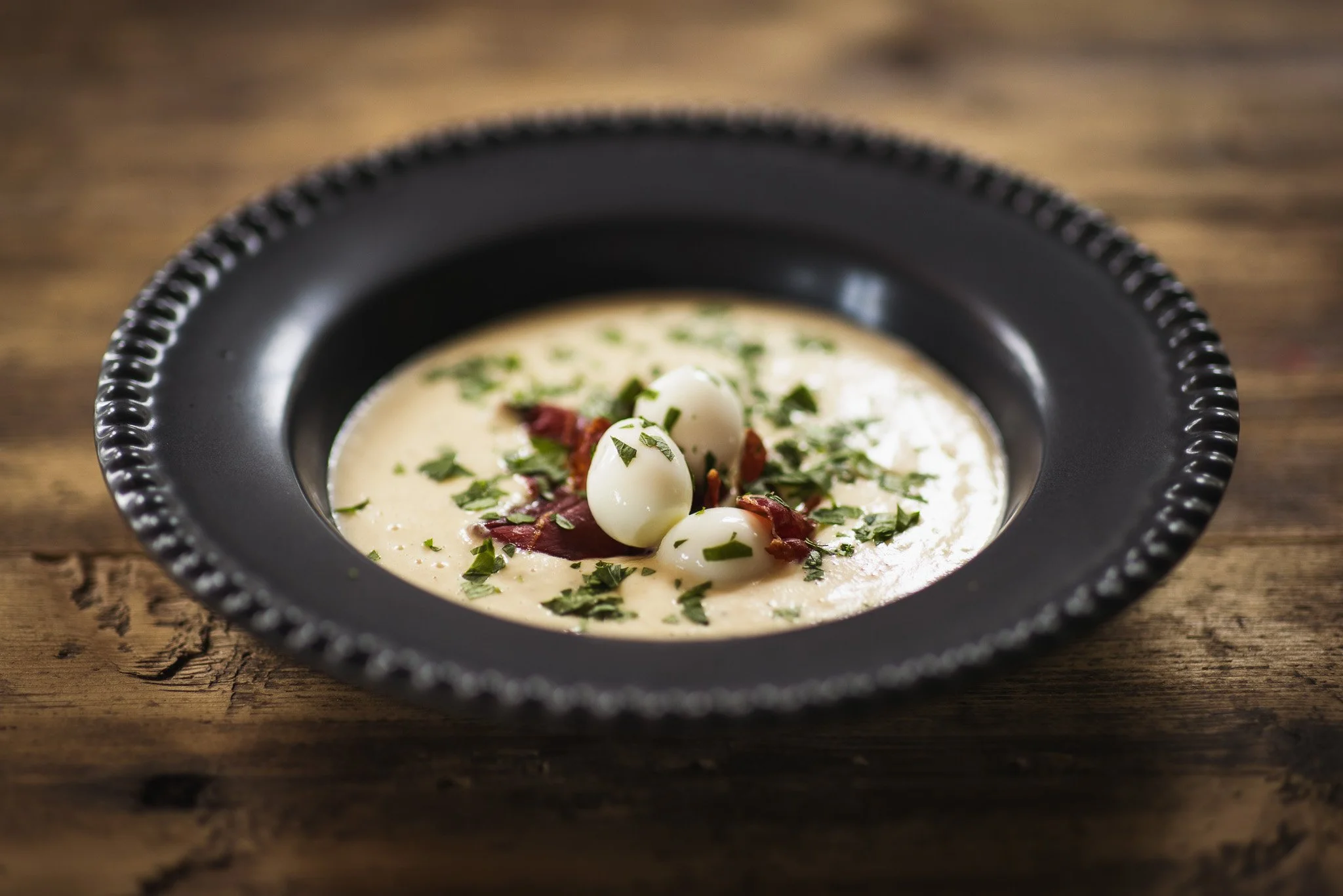 A bowl of creamy soup garnished with chopped herbs, quail eggs, and pieces of meat, served in a black dish on a rustic wooden table.