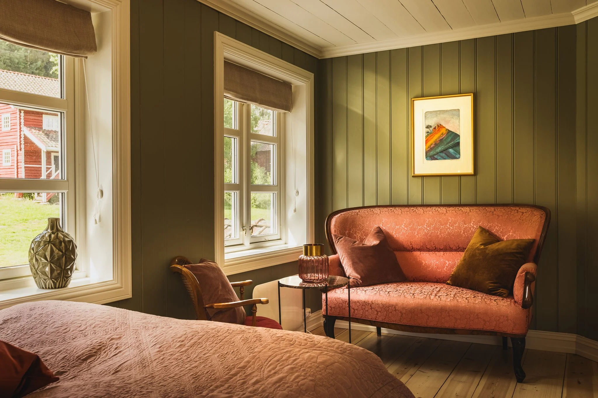 A cozy living room with green paneled walls, large windows with beige curtains, and a pinkish-orange vintage sofa with cushions. A framed abstract painting hangs on the wall, and a glass table with a pink vase is in front of the sofa.