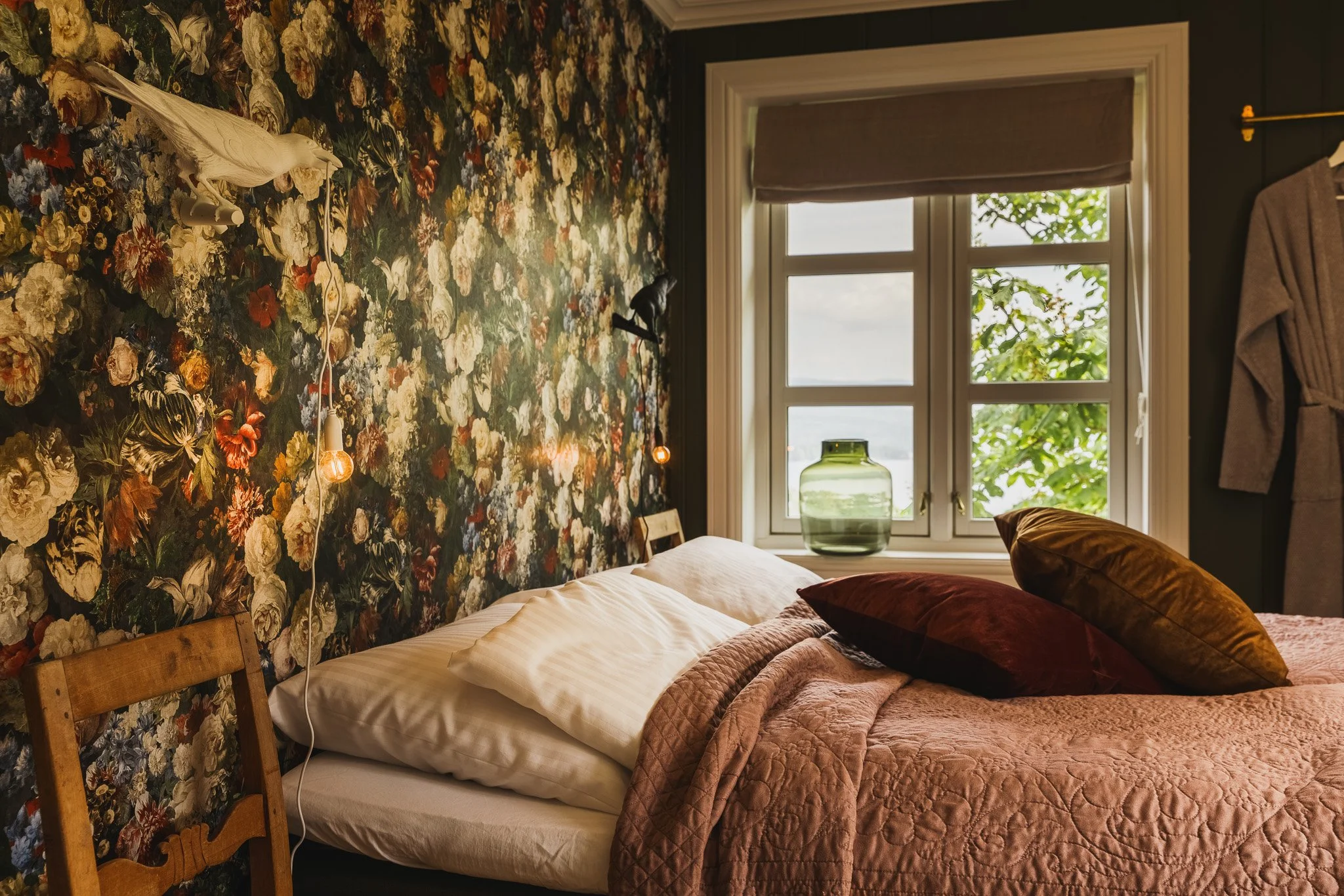 A bedroom with floral wallpaper on one wall, a wooden bed frame, and a window with a beige Roman shade. The bed has beige and brown pillows and a quilted pinkish comforter. There is a green glass vase on the window sill and some hanging light bulbs. 