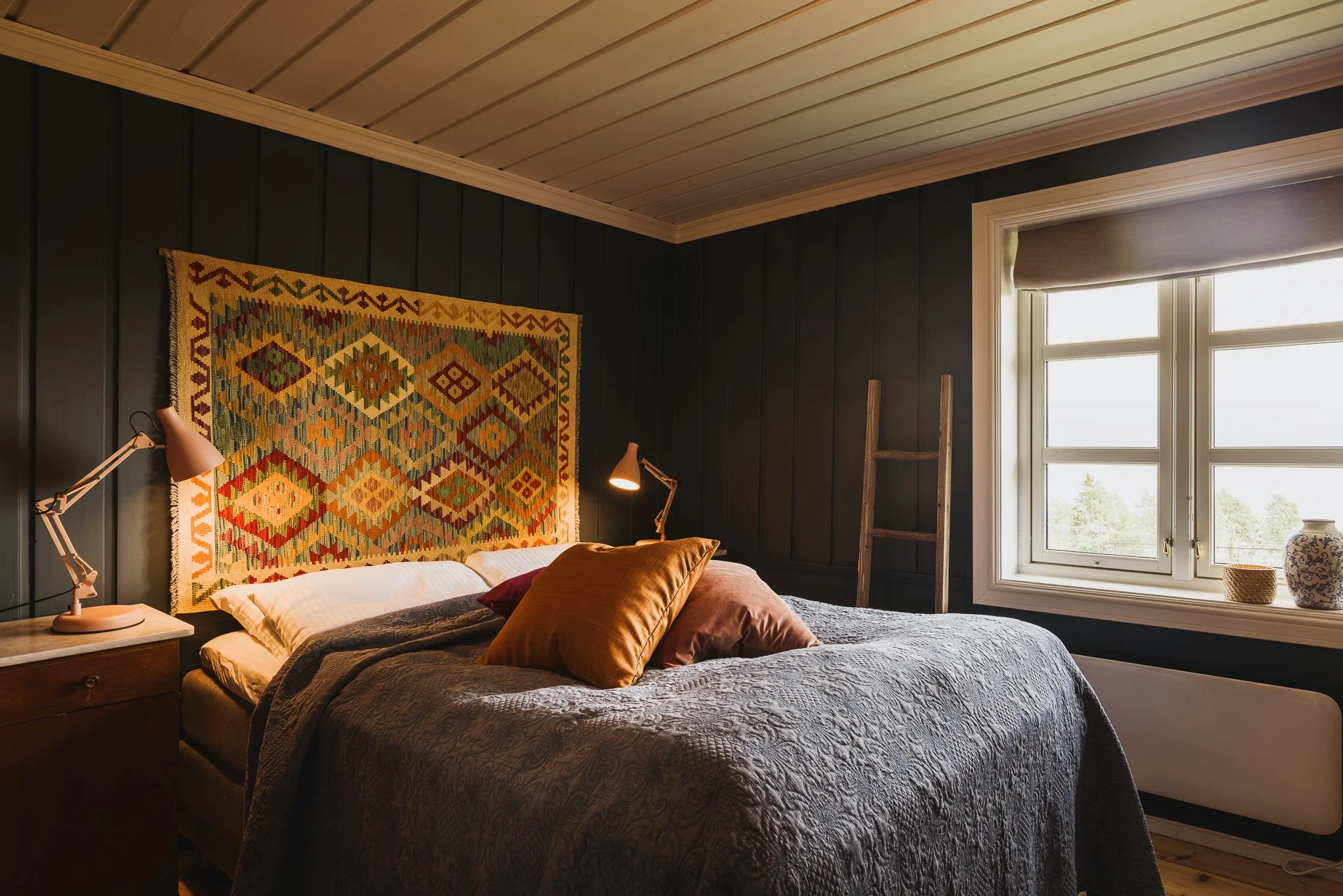 A cozy bedroom with dark paneled walls, a bed with pillows and a quilt, a bedside lamp, a ladder, a window with a view outside, and decorative items on the windowsill.