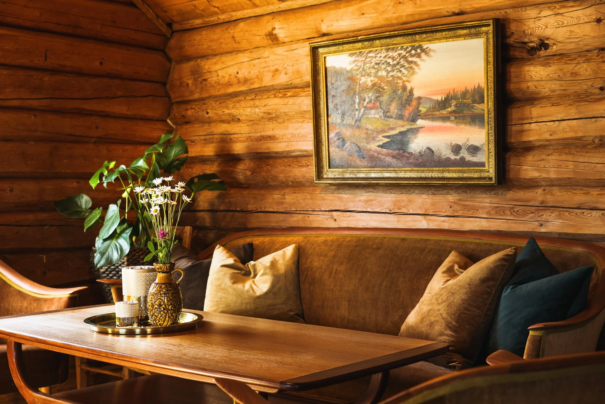 Interior of a cozy room with wood-paneled walls, a framed landscape painting, a wooden table with decorative vases, and a plant with white flowers and large green leaves.
