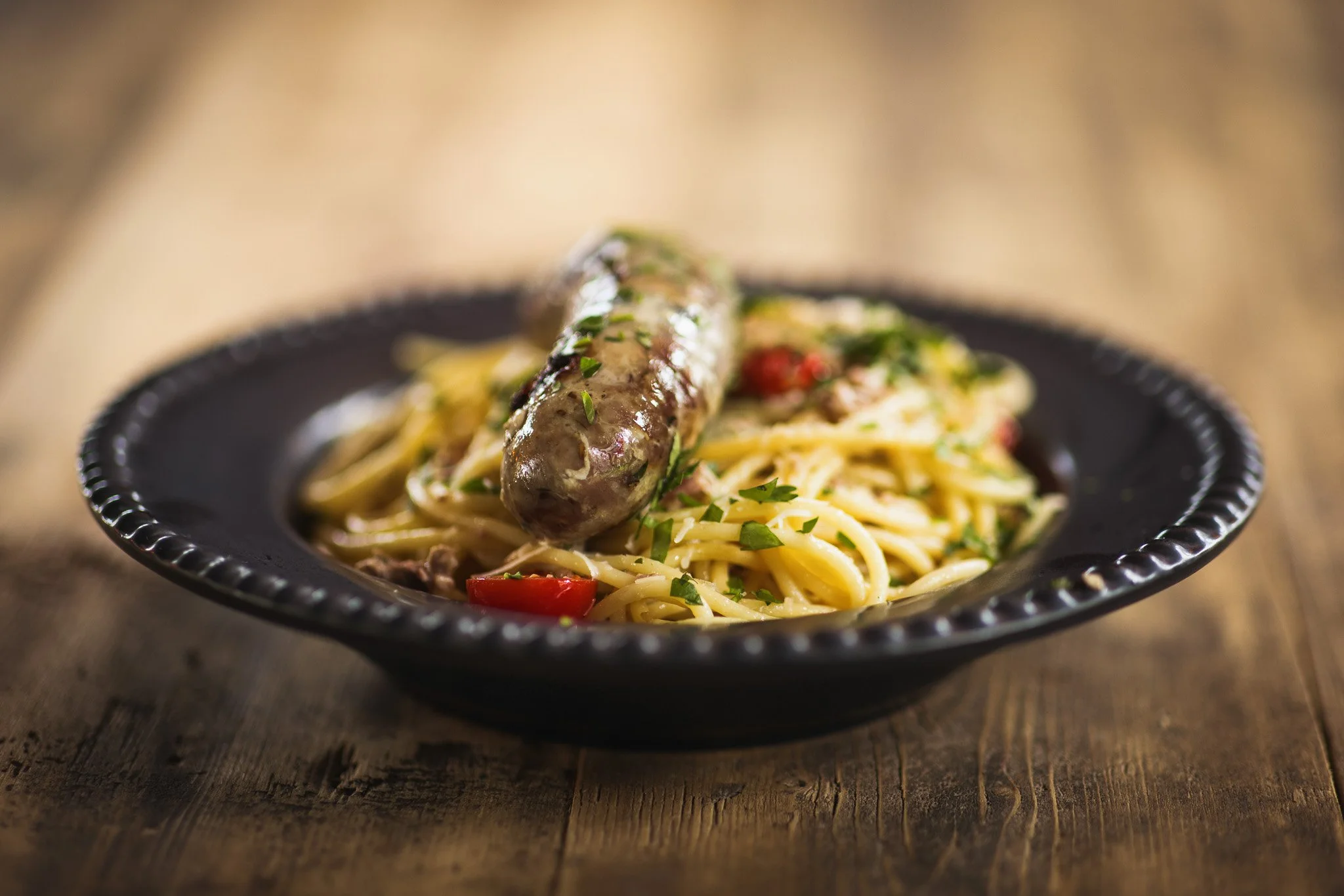 A plate of spaghetti with a sausage, garnished with chopped parsley and red chili peppers