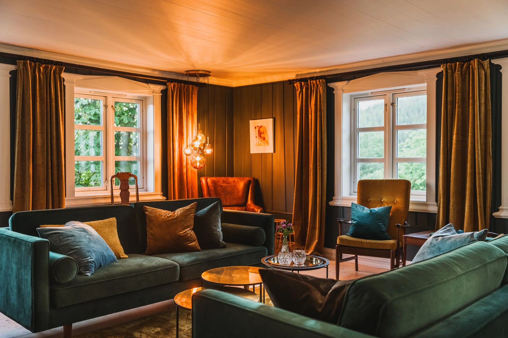 A cozy living room with dark green sofas and assorted pillows, wooden coffee tables, large windows with white trim and gold curtains, warm lighting from a hanging fixture, and a framed picture on a dark paneled wall.