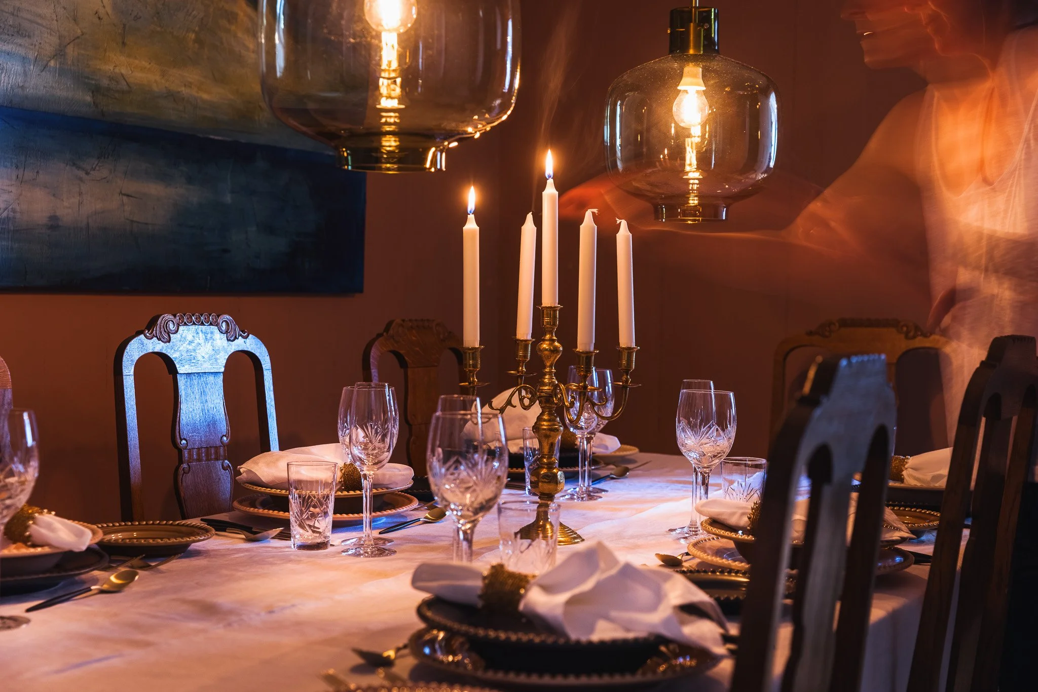 A dining table set for a formal dinner with white tablecloth, glassware, and gold-rimmed plates. Gold candelabra with white candles and large glass lanterns with Edison bulbs hanging from the ceiling illuminate the scene. Wooden chairs with carved ba