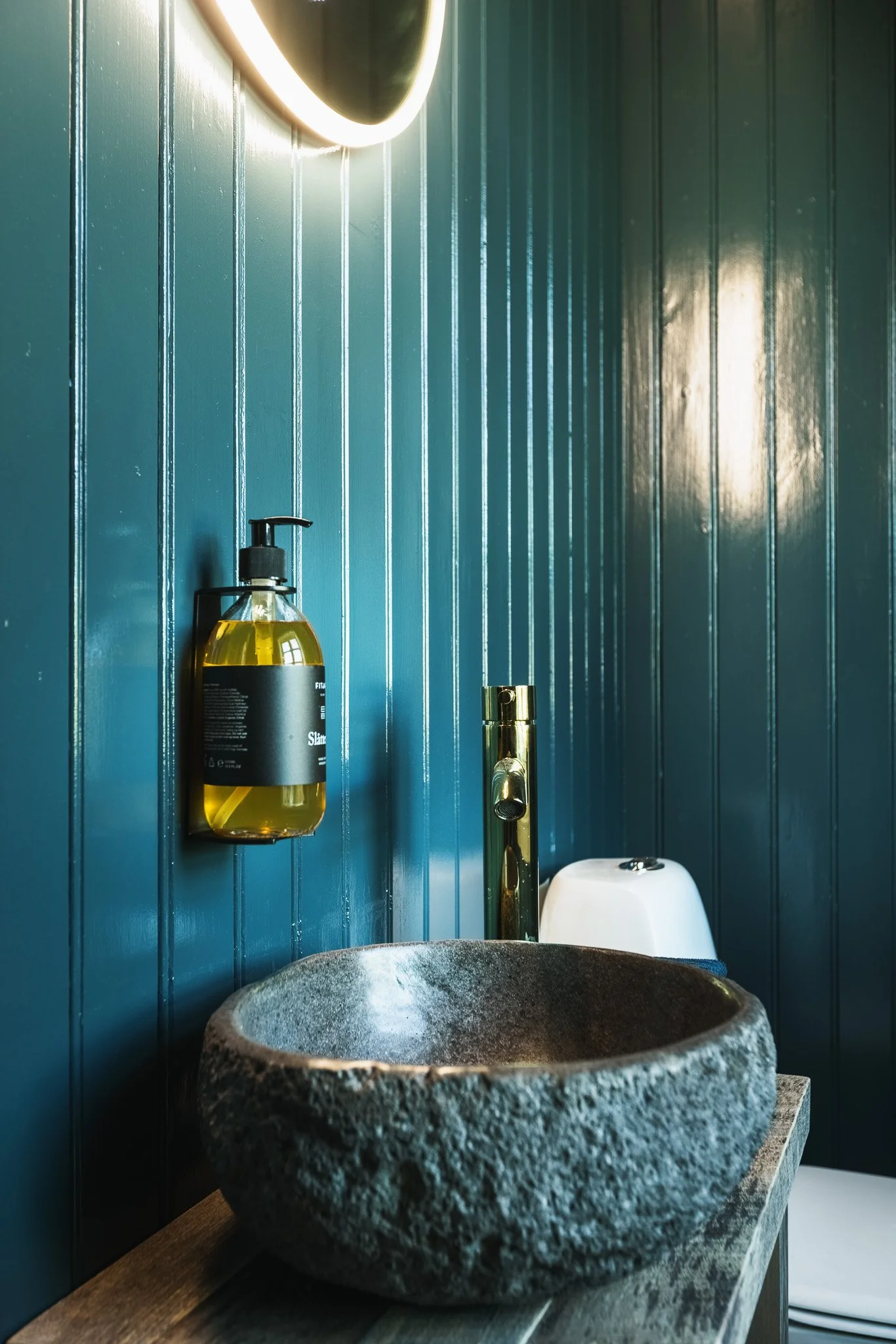 A rustic stone sink on a wooden countertop in a modern bathroom with blue vertical paneled walls, a gold faucet, and a soap dispenser with yellow liquid.