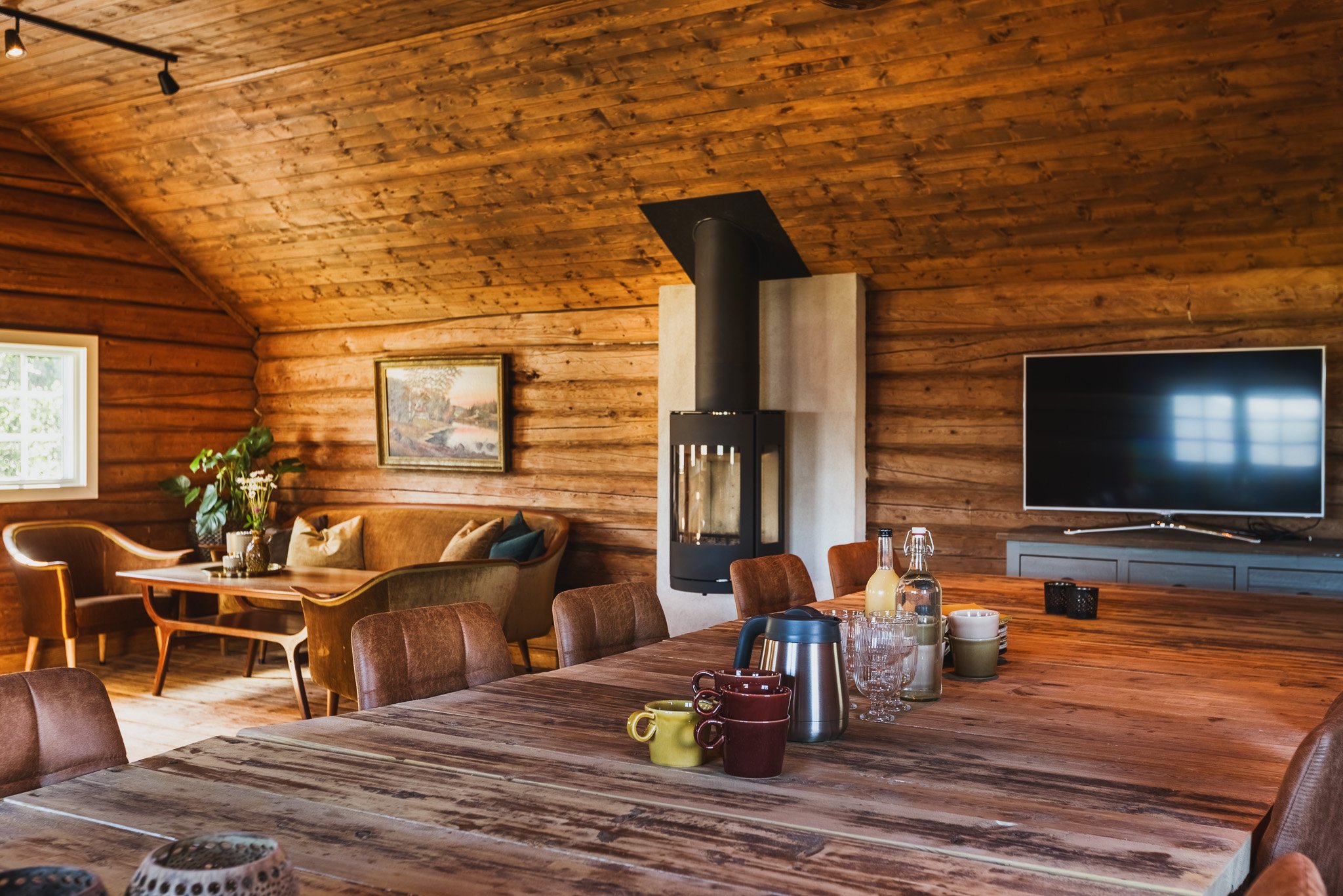 A cozy living room with wood-paneled walls and ceiling, a wood-burning stove, a flat-screen TV, a large wooden dining table with mugs, bottles, and glassware, and a seating area with a painting, plants, and comfortable chairs.