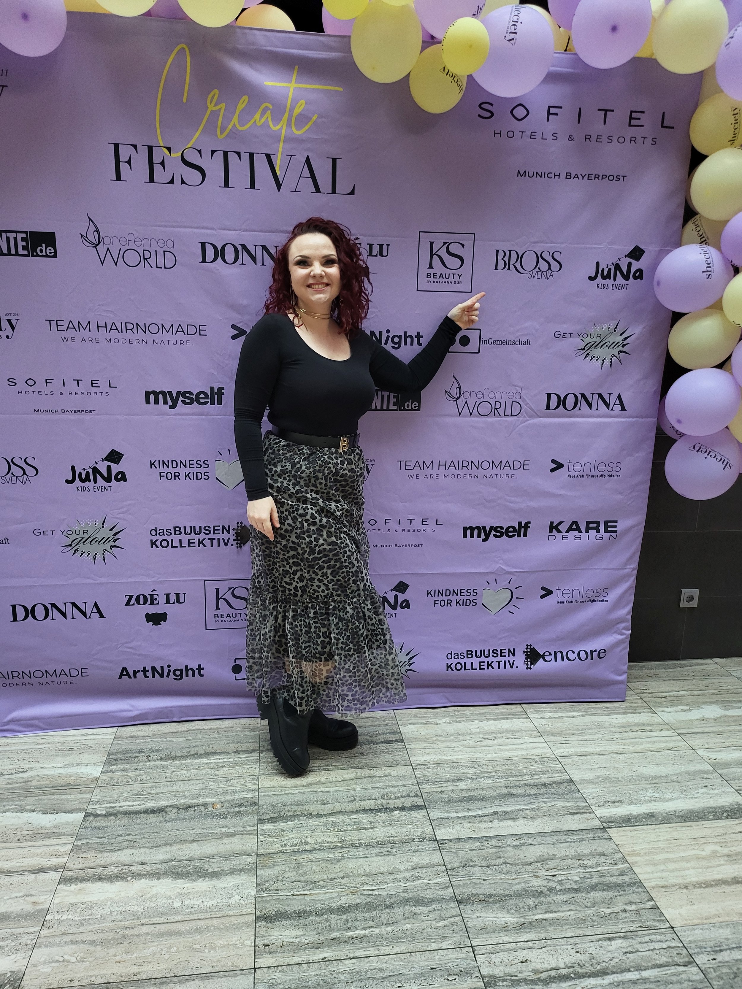 Eine Frau mit dunkelroten, gelockten Haaren trägt eine schwarze langärmlige Oberbekleidung und eine Leopardenhose. Sie steht vor einer lilafarbenen Festival-Banner, das mit Ballons dekoriert ist, und zeigt auf ein Logo auf dem Banner.