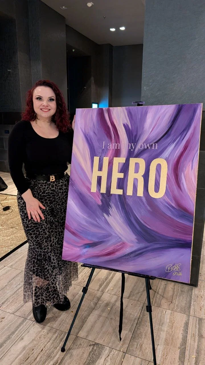 Eine Frau mit lockigem roten Haar steht neben einem großen, bunten Gemälde auf einem Ständer. Das Gemälde hat einen abstrakten Hintergrund in Violett- und Rosatönen mit dem Text: „I am my own HERO“. Die Frau trägt eine schwarze Oberbekleidung, eine Leopardenhose und schwarze Schuhe.