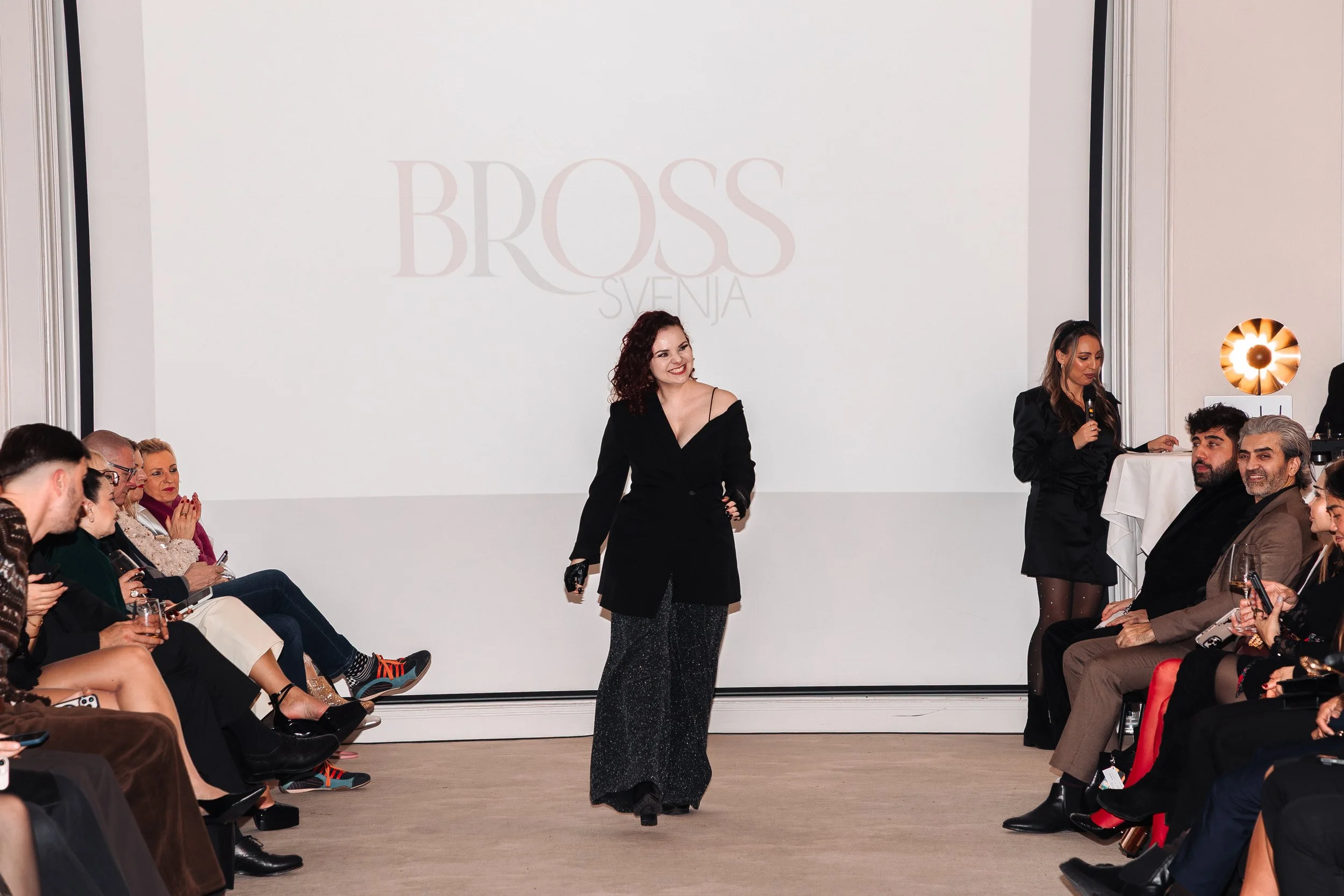 Moderner Laufsteg mit Model, Publikum und großem Bildschirm im Hintergrund. Die Zuschauer sitzen an beiden Seiten, der Moderator hält ein Mikrofon, auf dem Bildschirm steht 'BOSS SVEHNIA'.