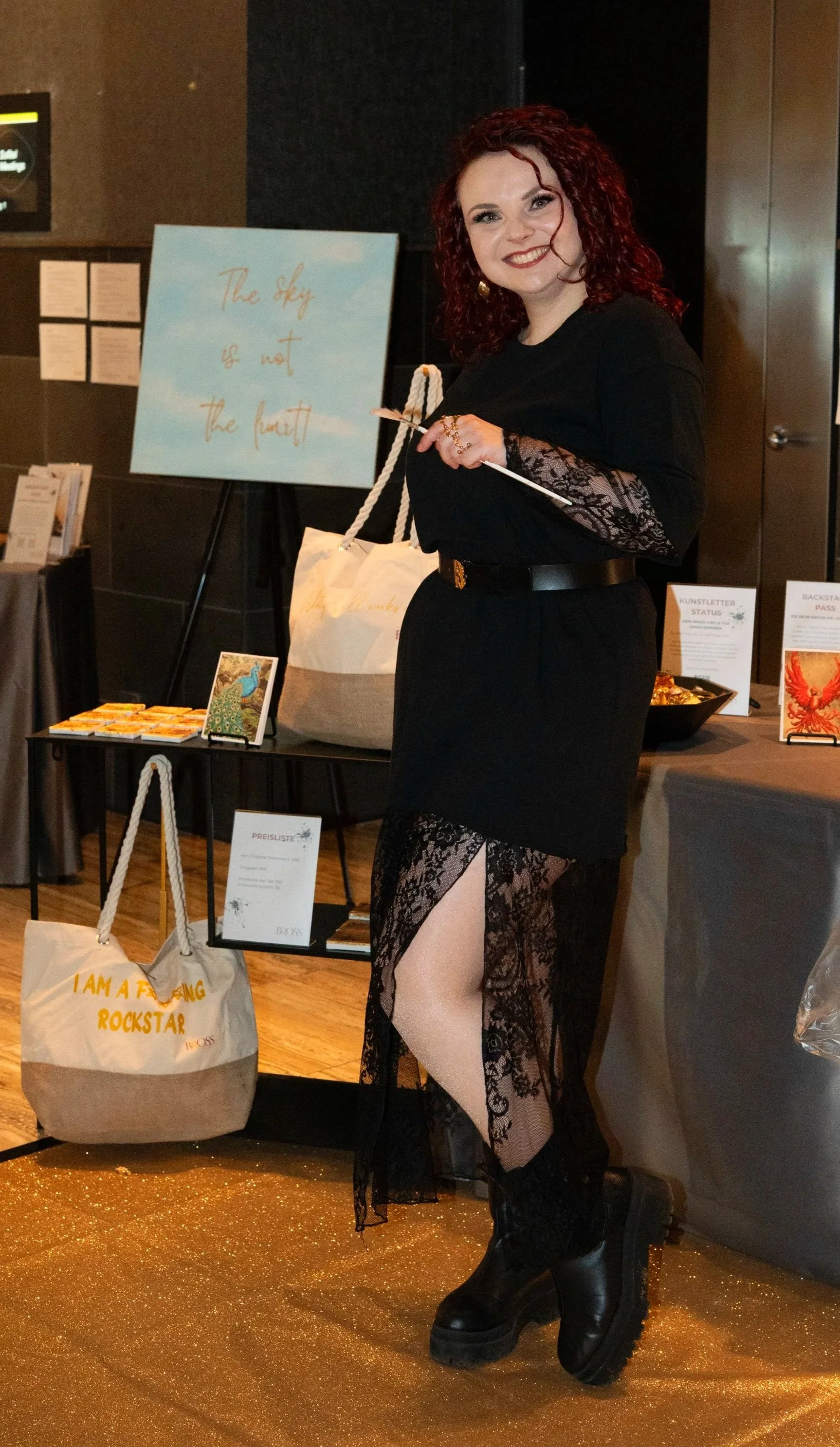 Frau in schwarzem Kleid mit Spitze und hohen Stiefeln steht vor einem Tisch mit Broschüren und Taschen, hinter ihr ein Plakat mit dem Text „The sky is not the limit“.