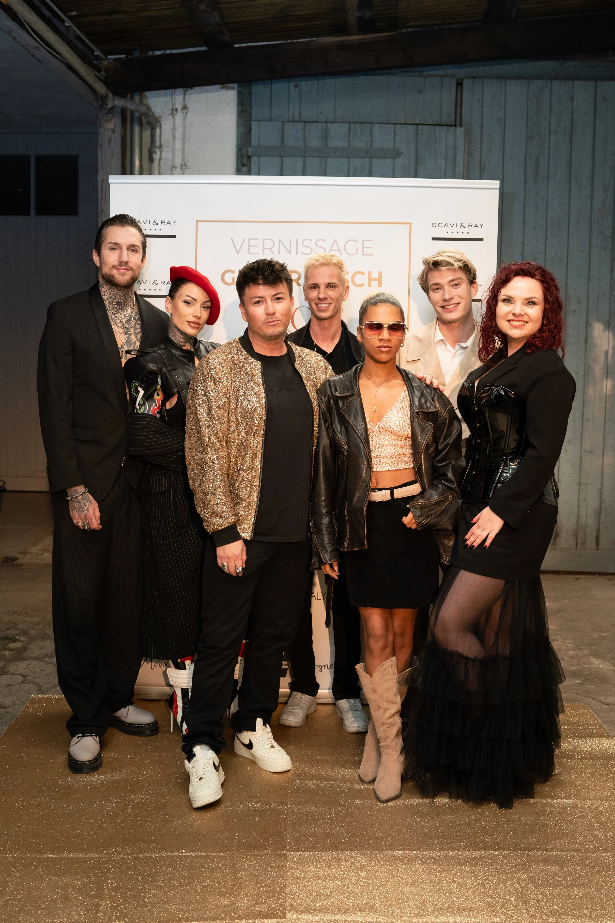 Eine Gruppe von acht Menschen poseiert vor einem Banner mit der Aufschrift 'Vernissage GURU GÖCH' bei einer Veranstaltung. Sie tragen moderne, stylische Kleidung, einige mit auffälligem Make-up und Accessoires.
