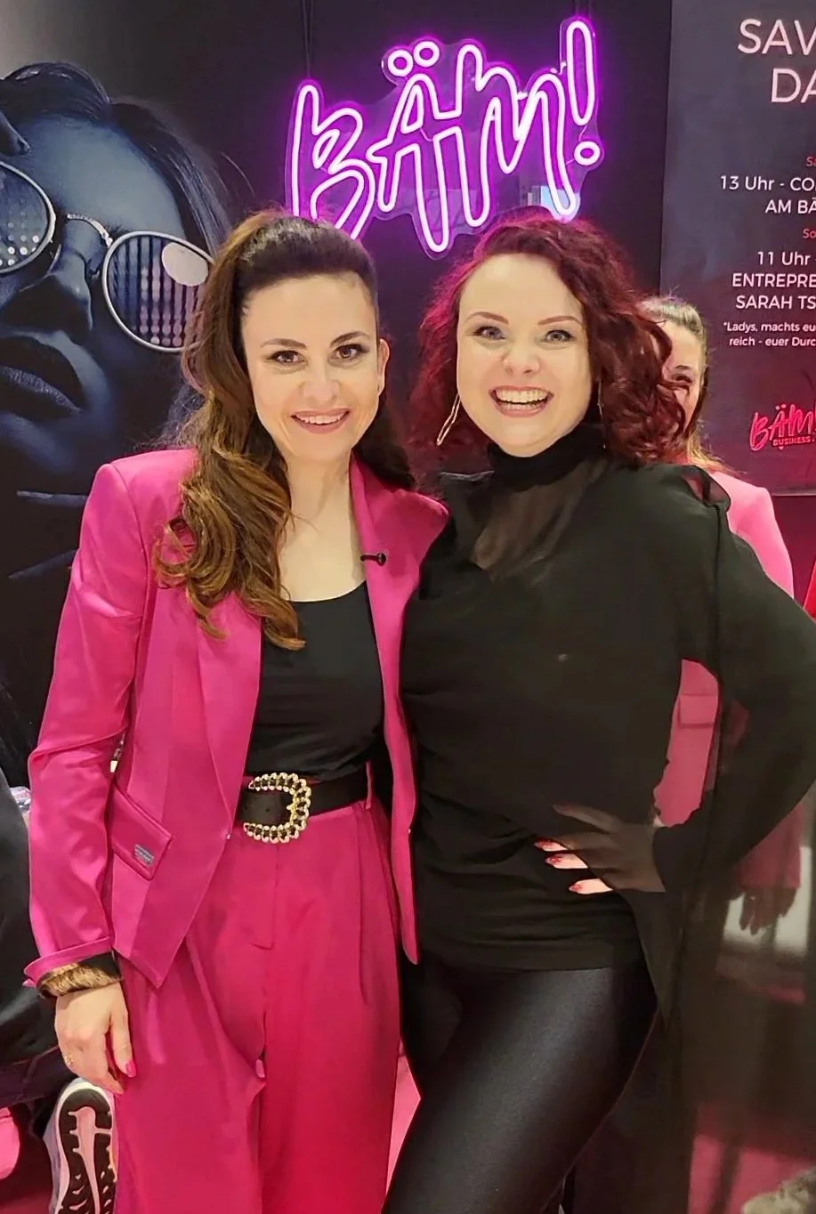 Zwei lachende Frauen stehen nebeneinander vor einem schwarzen Hintergrund mit einer großen neonlila Schrift, die 'BÄM!' sagt, und einem Poster mit einem schwarzen und weißen Porträt einer Frau mit Sonnenbrille.