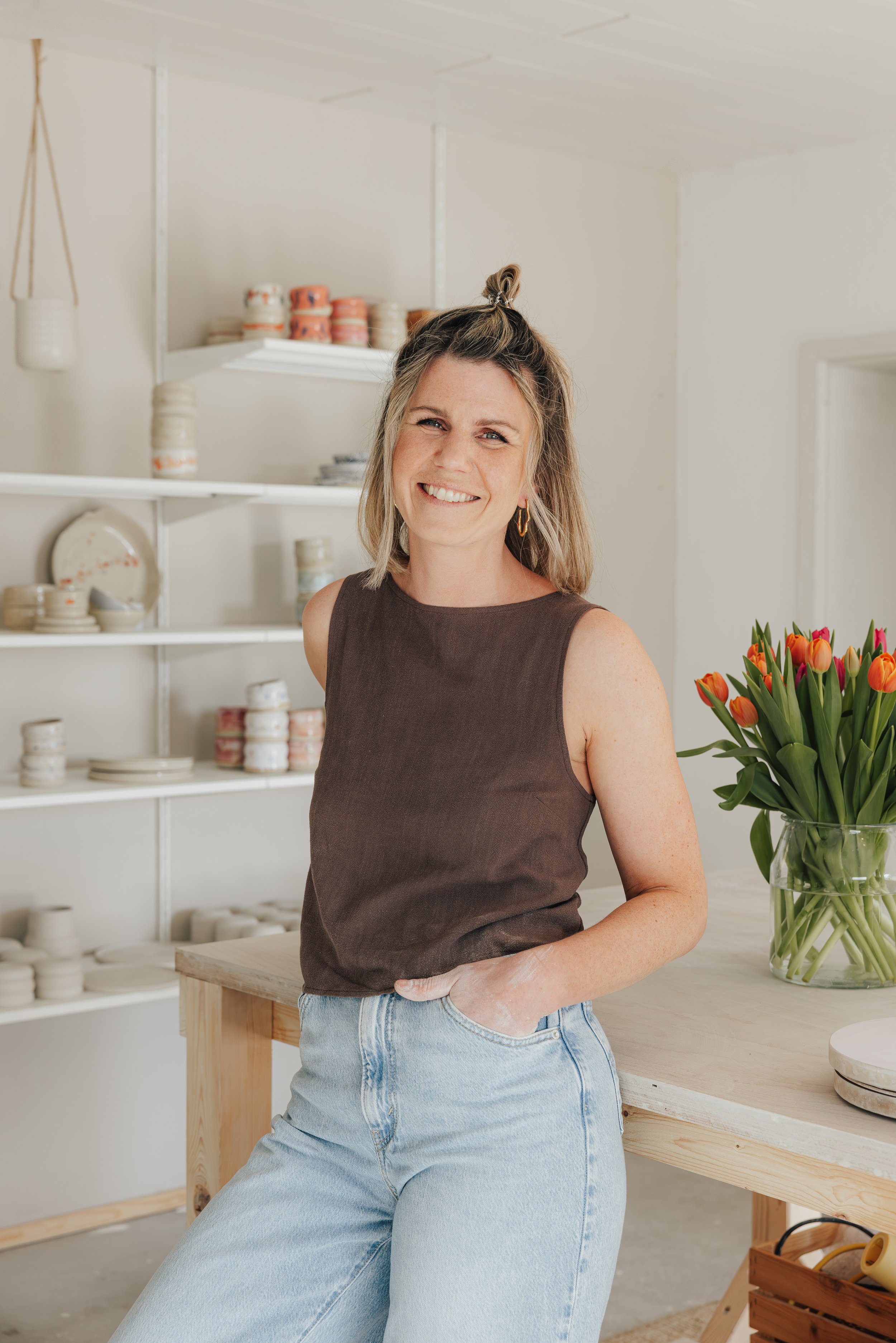 Eine lächelnde Frau steht in einer hellen, minimalistischen Küche mit weißen Regalen, Keramikgeschirr und einem großen Blumenstrauß aus Tulpen auf dem Tisch.