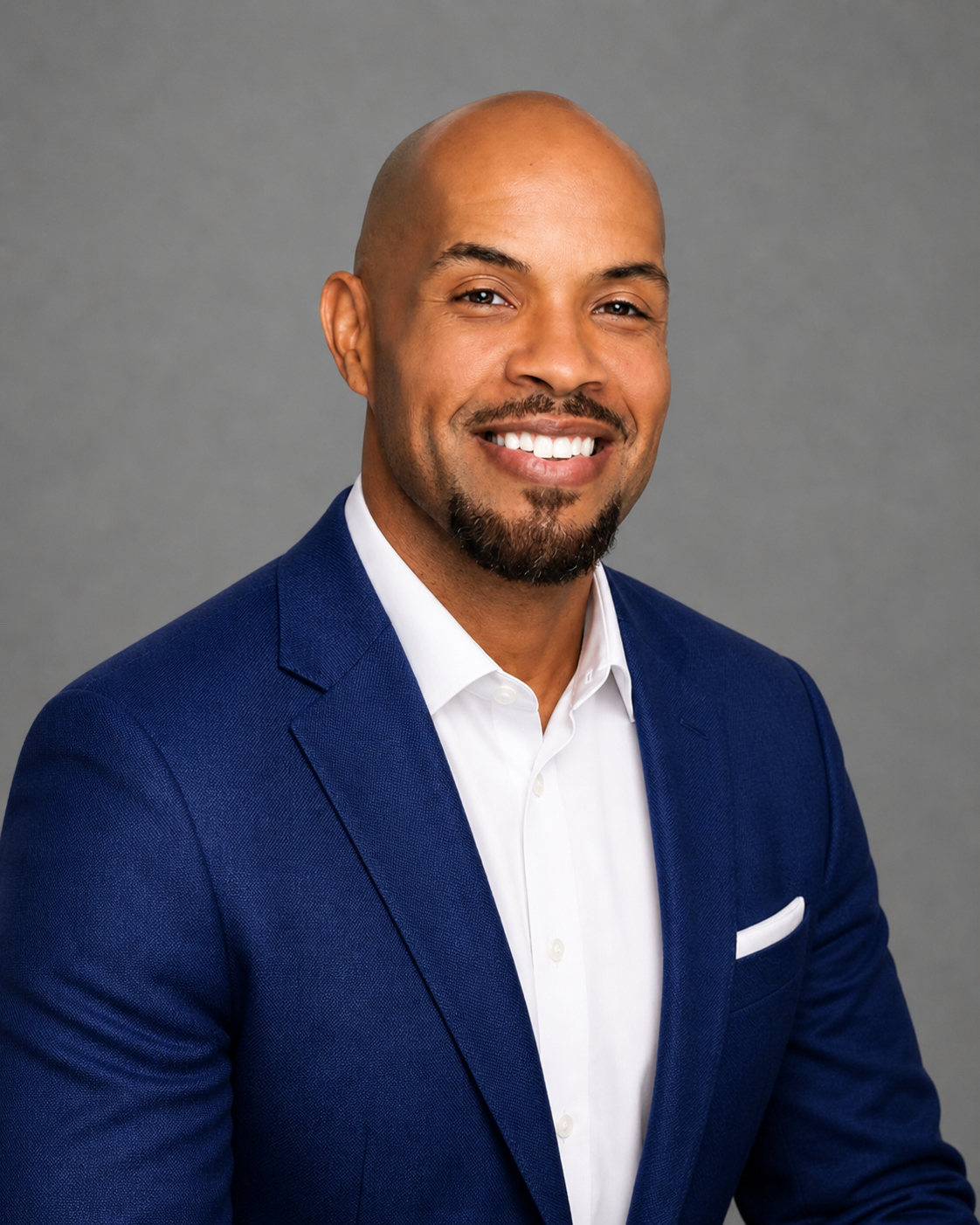 Headshot of a bald man with a goatee, wearing a white shirt and blue suit, smiling against a gray background.