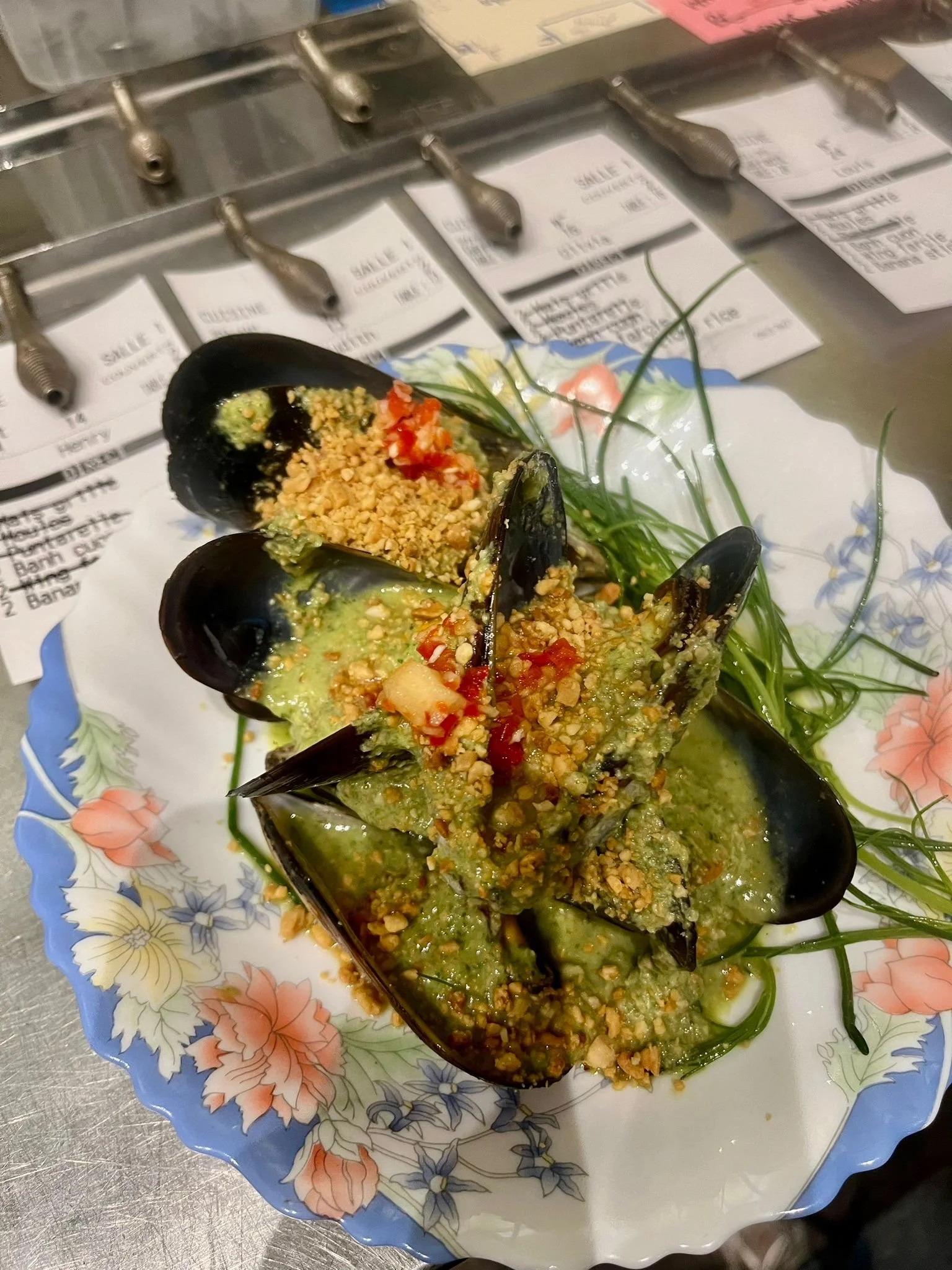 Baked mussels topped with crushed peanuts, diced red peppers, and green sauce, garnished with fresh green herbs on a floral-patterned plate.