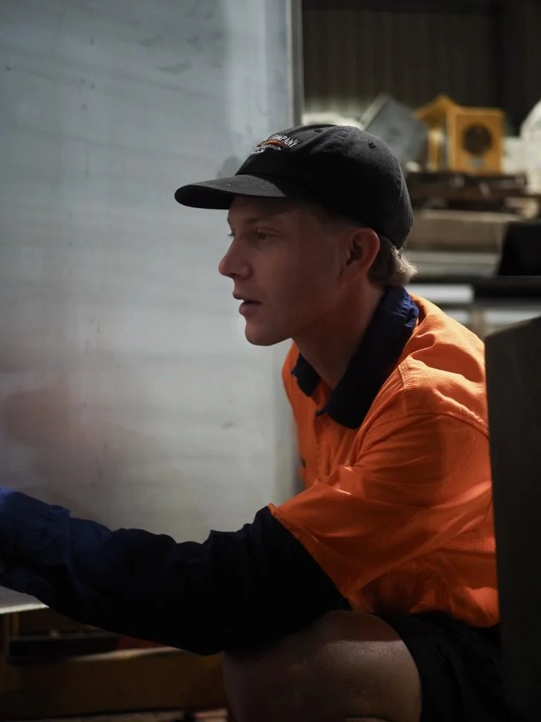 A young man wearing an orange work shirt, black cap, and black long sleeves, sitting indoors, looking to the left.